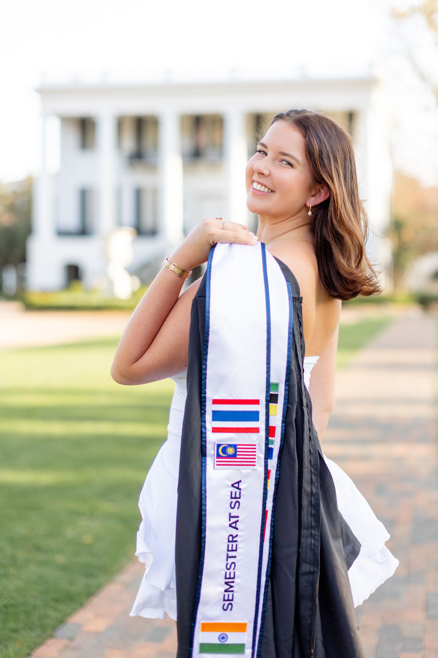 Student wearing graduation sash with international flags smiles while standing on campus lawn near white building.