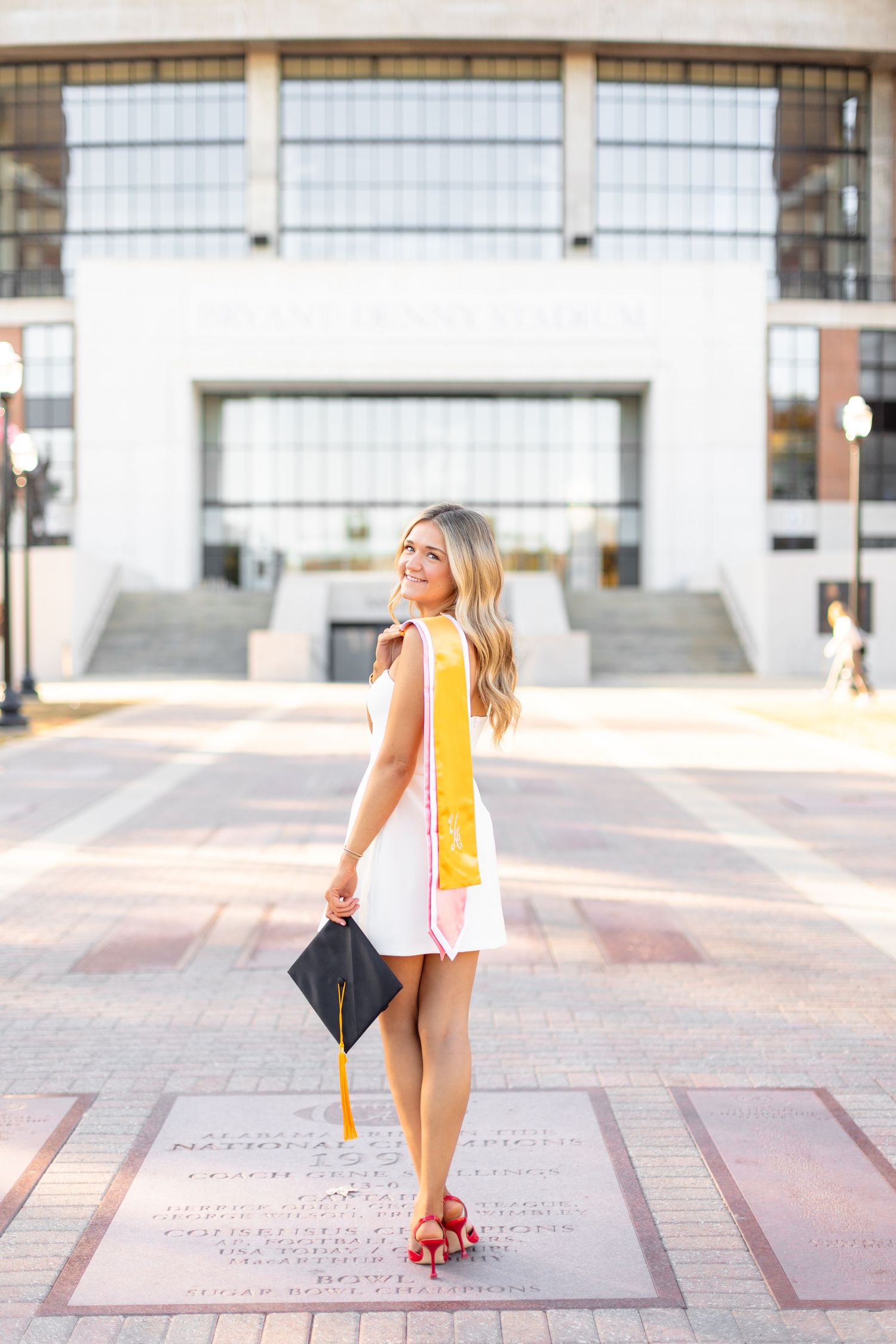 A graduate in a white dress and yellow stole poses on a campus plaza holding a graduation cap.