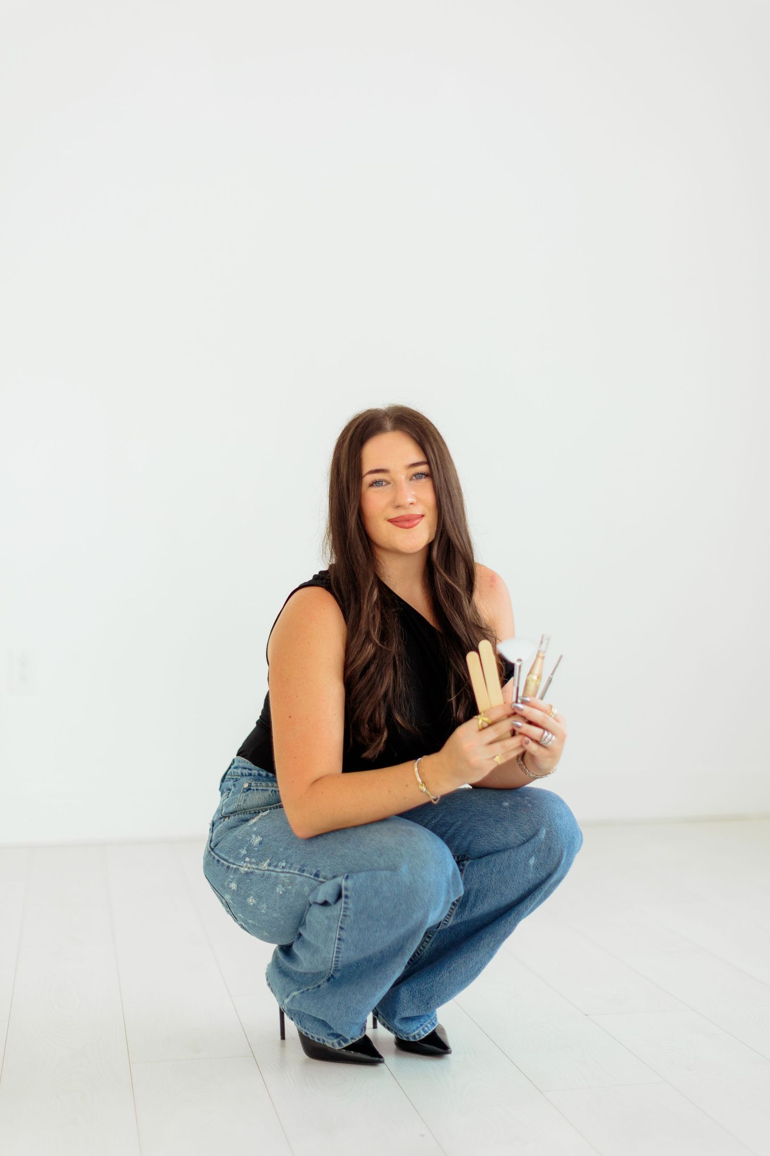 Young woman crouching in jeans and black top, holding makeup brushes, smiling against white background.