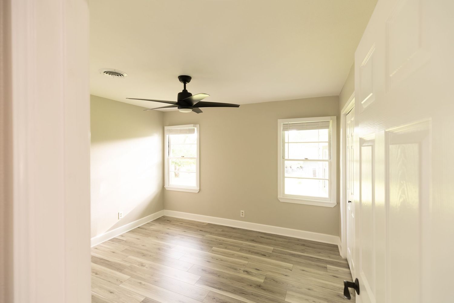 Bright empty bedroom with ceiling fan, two windows, light hardwood floors, and neutral beige walls.