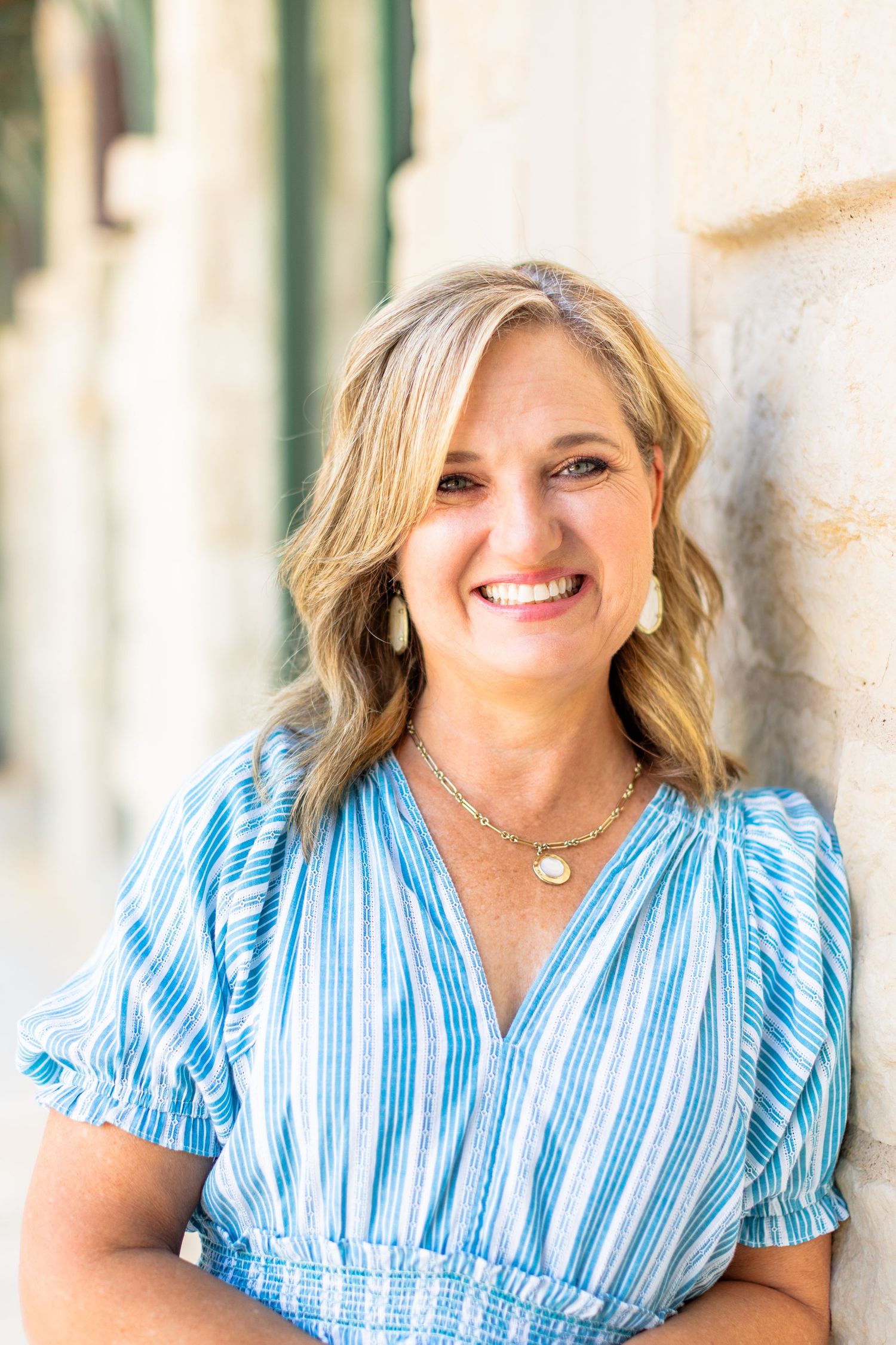 A person in a blue and white striped dress smiles while leaning against a wall outdoors.