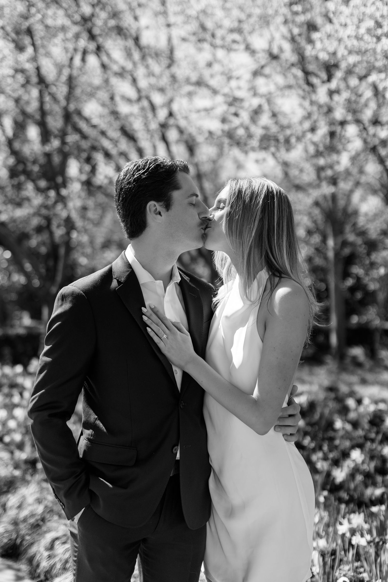 A romantic black and white portrait of a couple sharing a tender kiss in a garden setting with blooming trees.