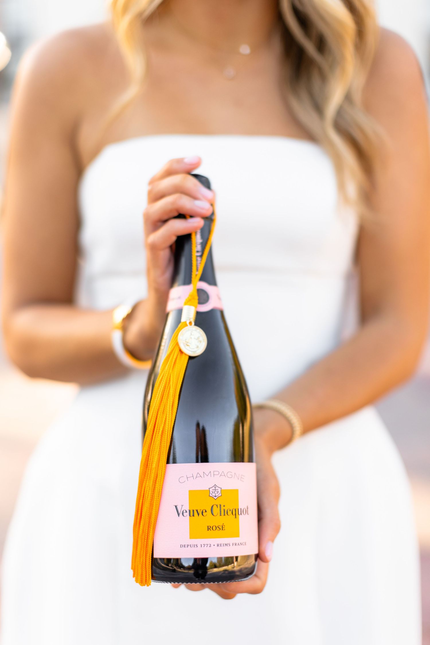 Hands holding a bottle of Veuve Clicquot champagne with gold tassel decoration against a white dress.