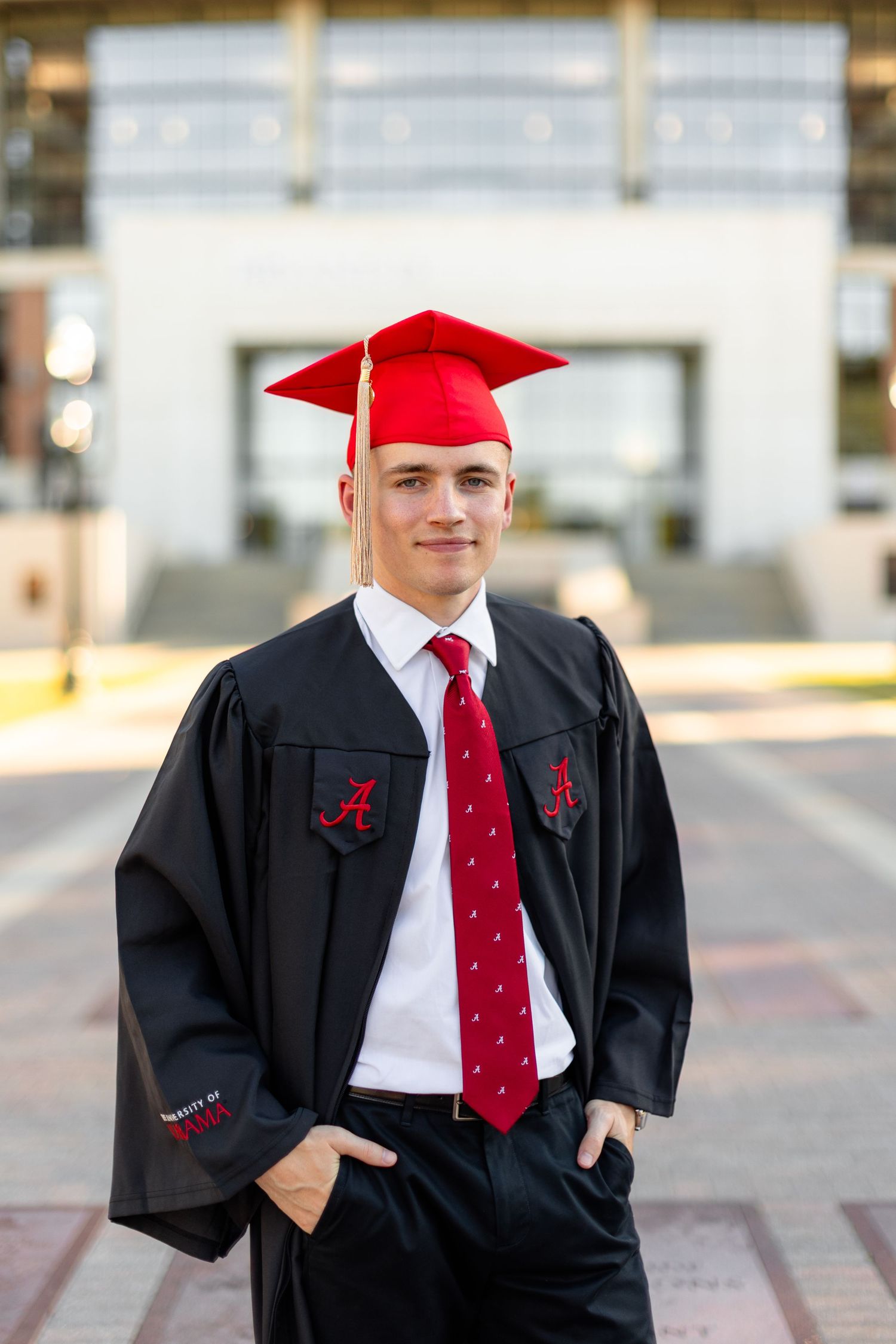 Graduate wearing red cap and black gown stands confidently in front of modern campus building.