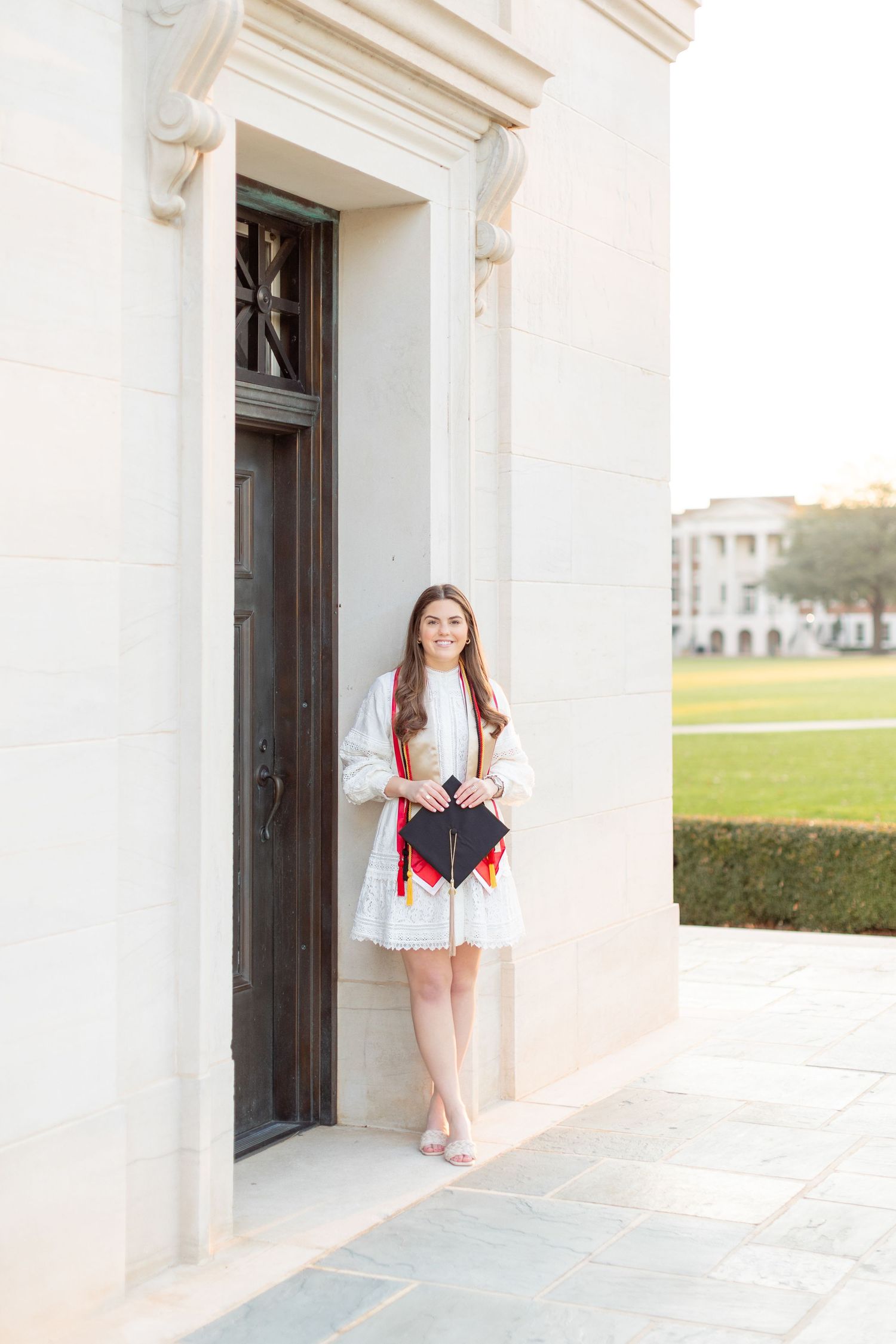 Graduate in white dress poses elegantly in front of classical architectural doorway.