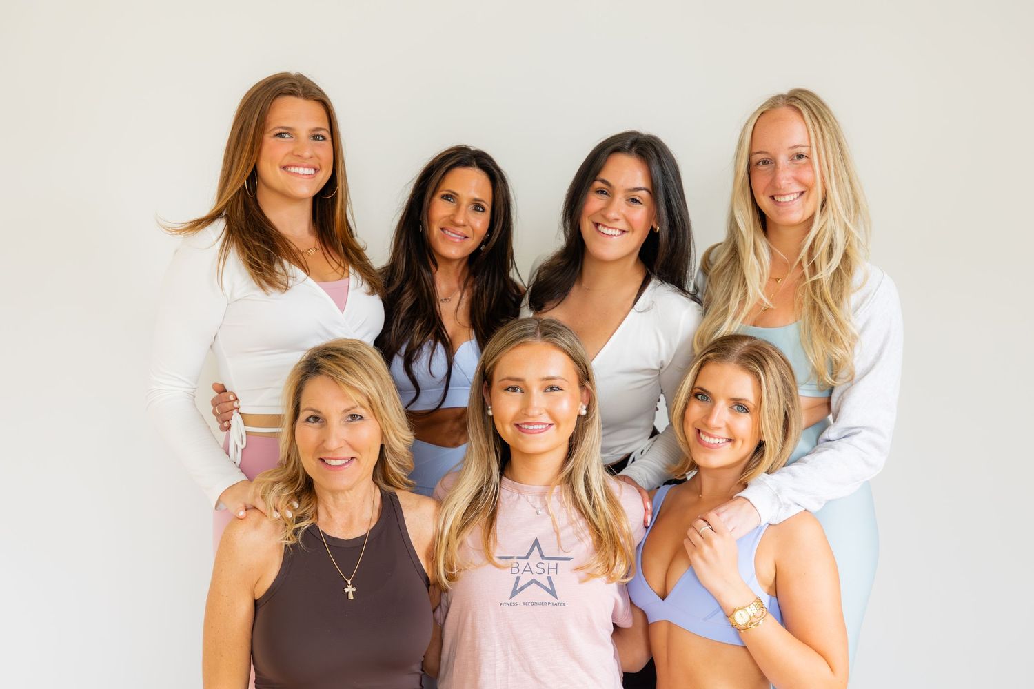 A group of seven women wearing white and neutral colored tops smile together in a casual portrait against a light background.