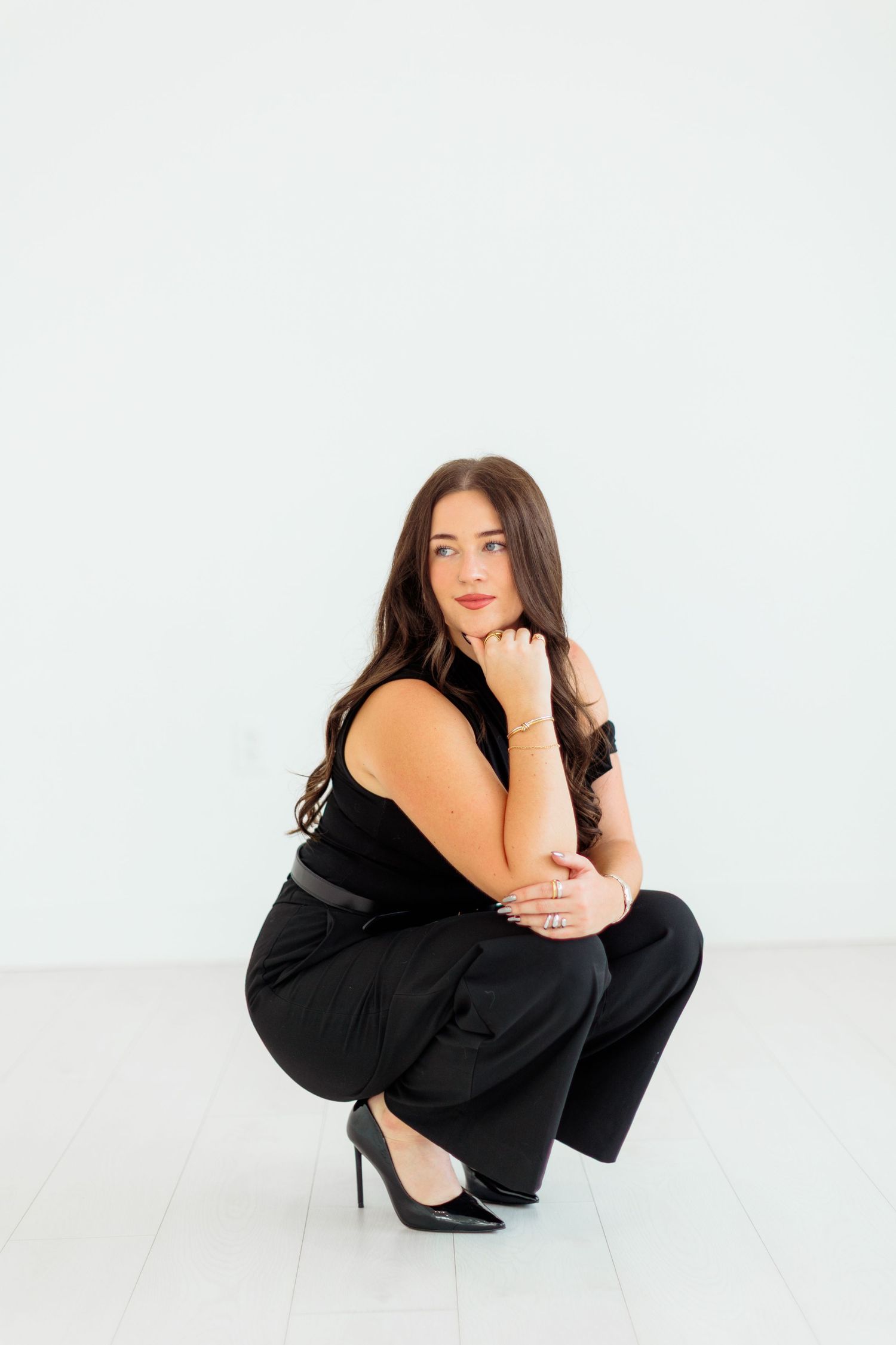 Woman in black outfit crouching gracefully in heels against white wall.