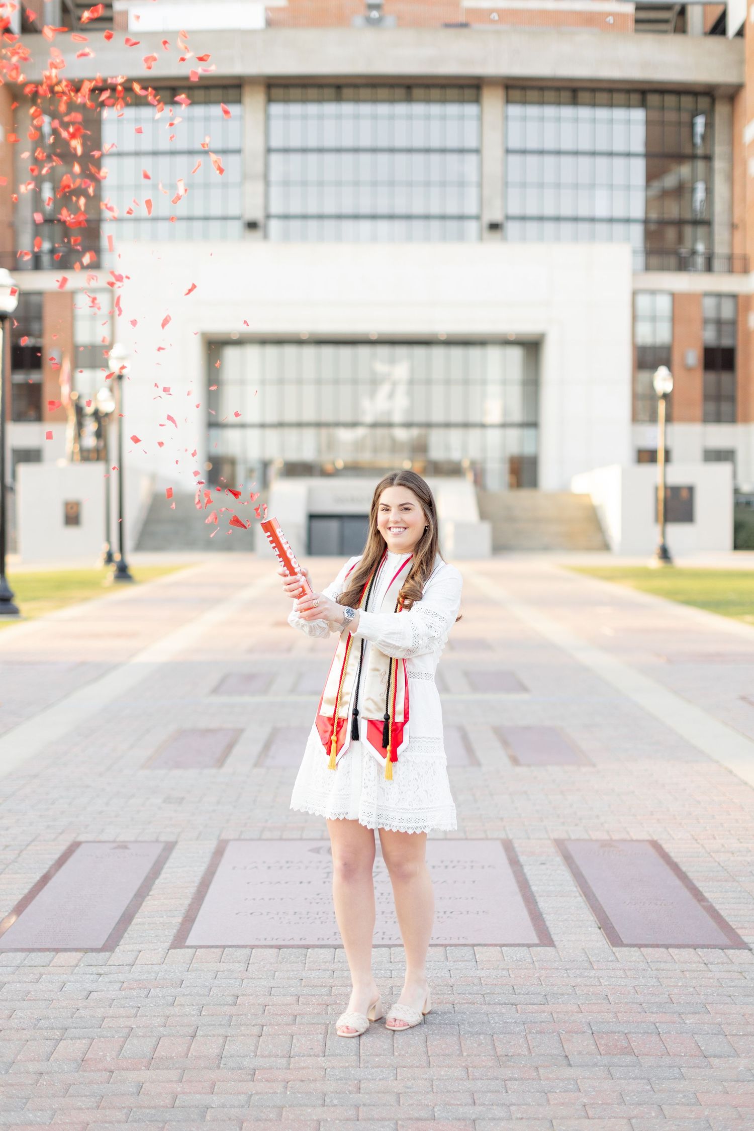 Graduate in white dress celebrates on brick plaza with red confetti popper in front of modern building.