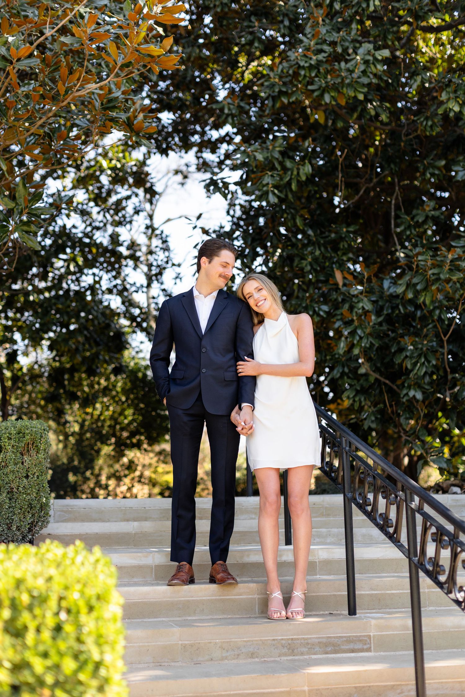 A couple stands together on concrete steps surrounded by lush greenery and trees in an outdoor setting.