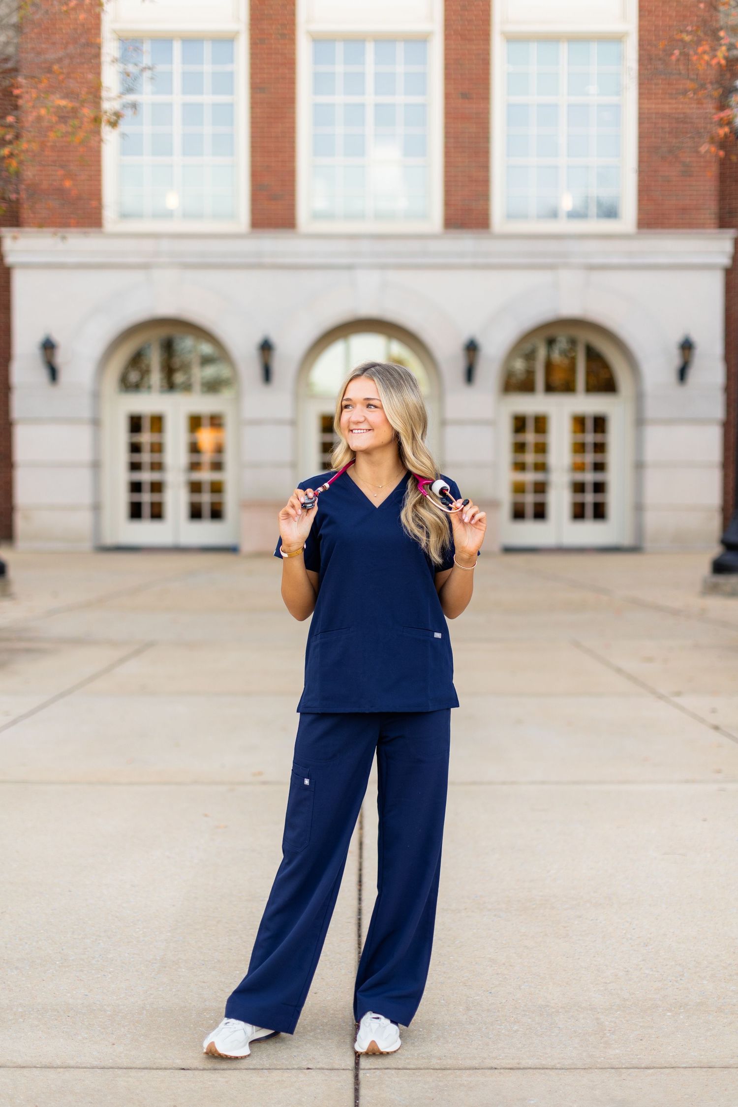 Healthcare professional in navy blue scrubs stands smiling in front of brick building with arched windows.