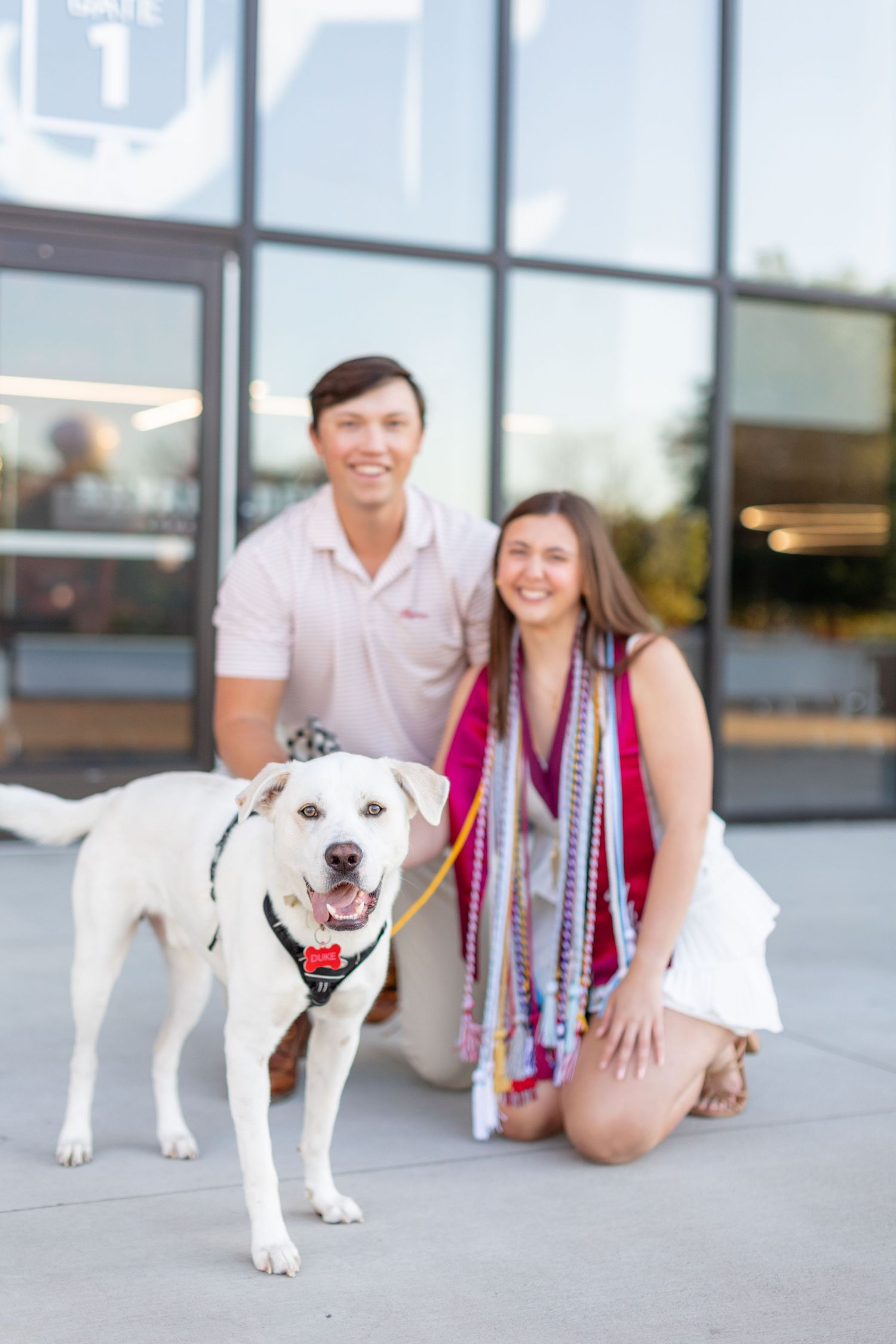 Graduates pose with white dog wearing red bandana outside glass building.