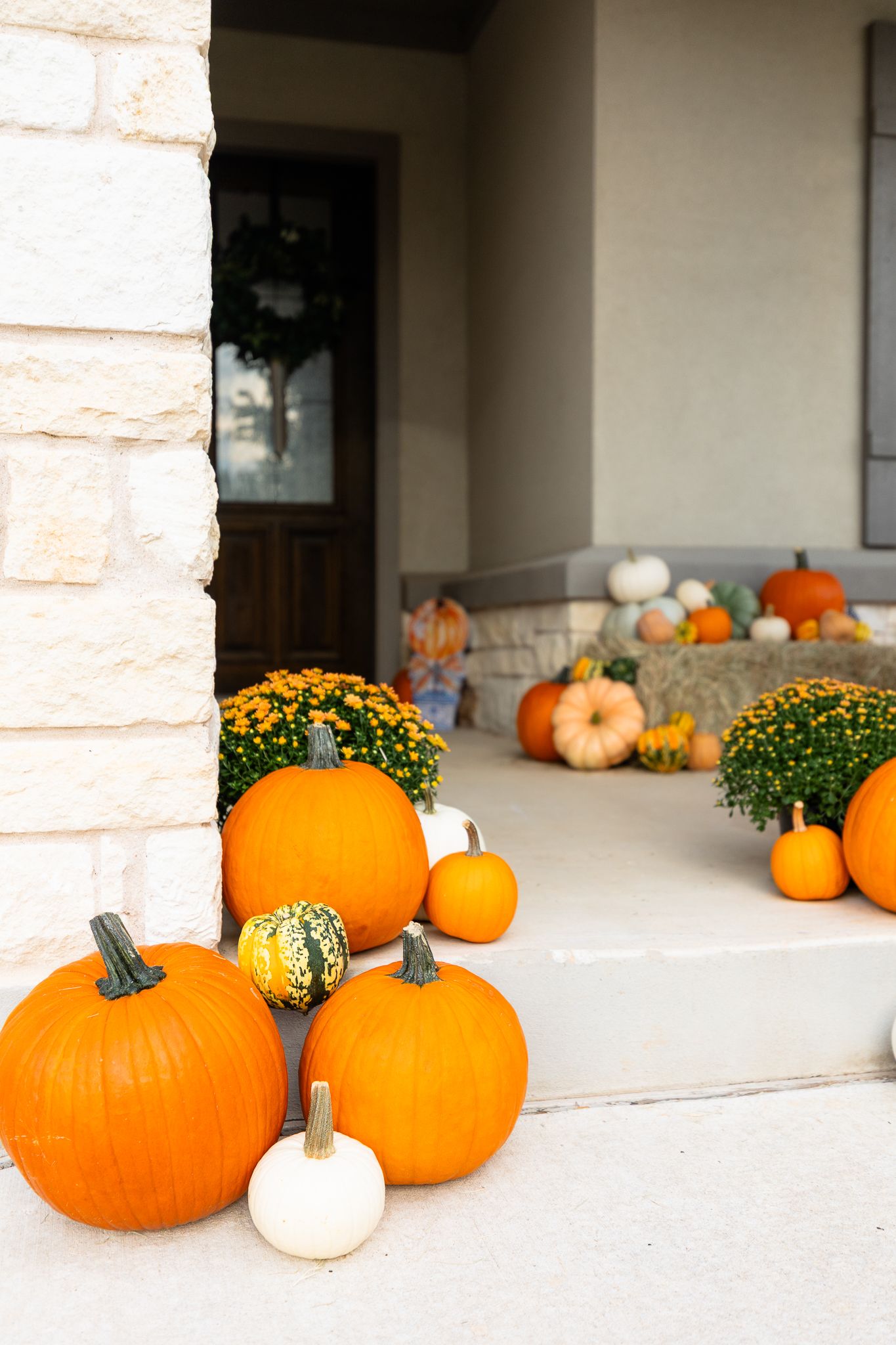 Orange pumpkins and yellow gourds are arranged decoratively along a stone porch with autumn mums.