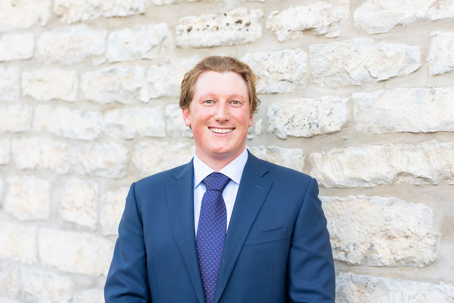 Professional headshot of a person wearing a navy suit and purple tie against a light stone wall background.