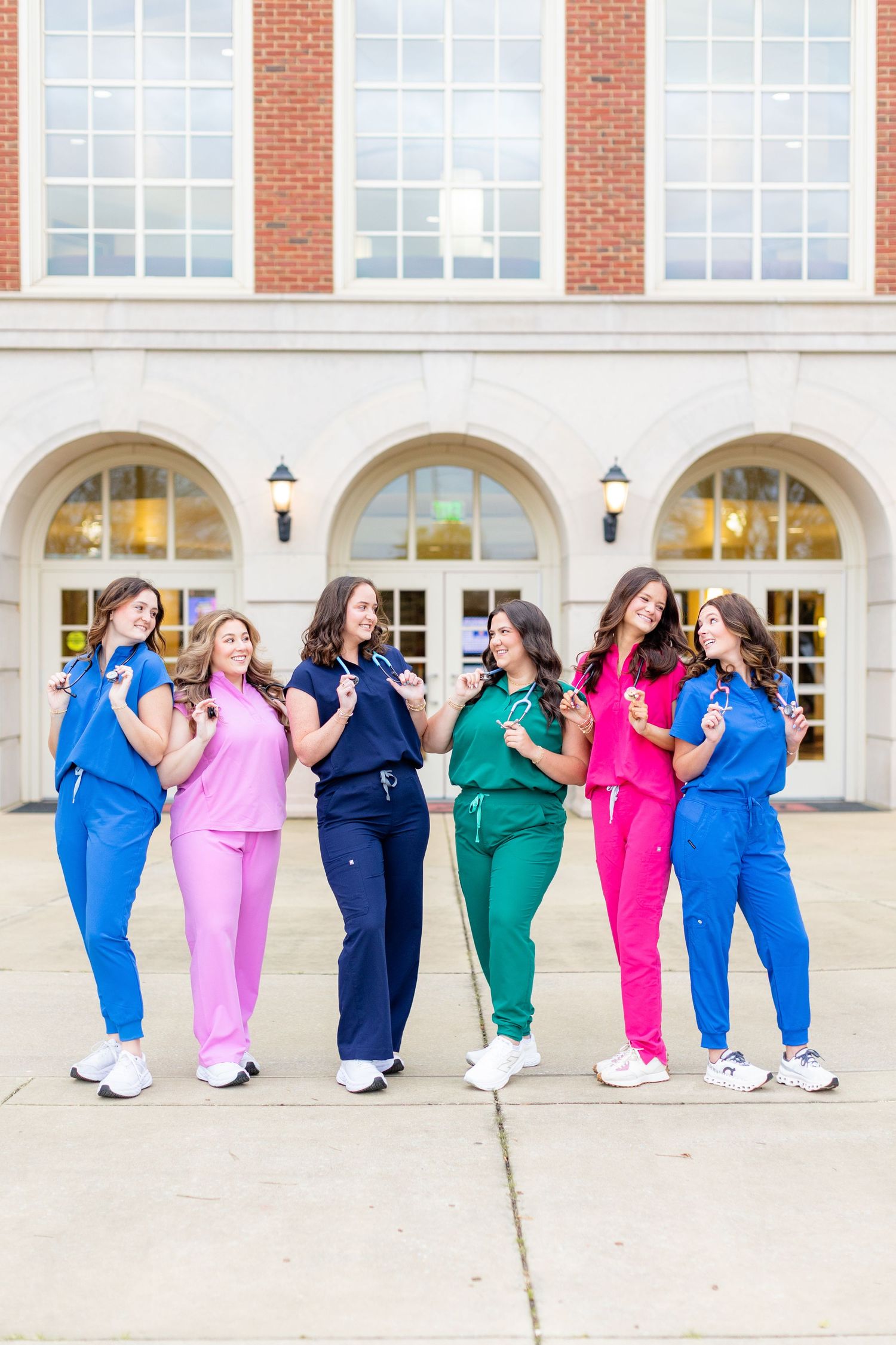 Medical professionals in vibrant colored scrubs pose playfully outside brick building with arched windows.