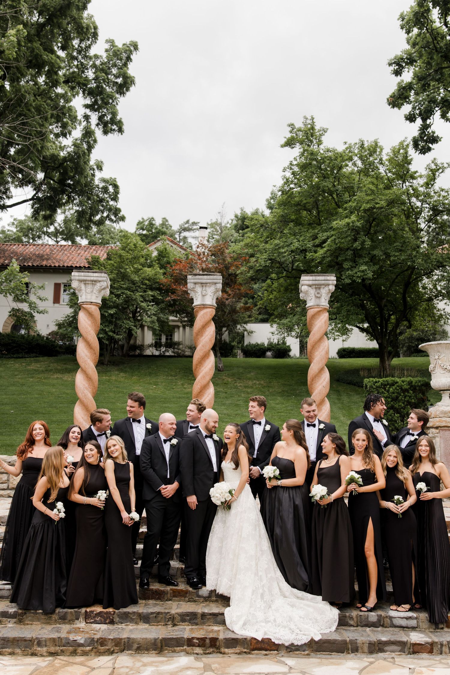Wedding party in black formal attire poses on stone steps with ornate spiral columns behind them.