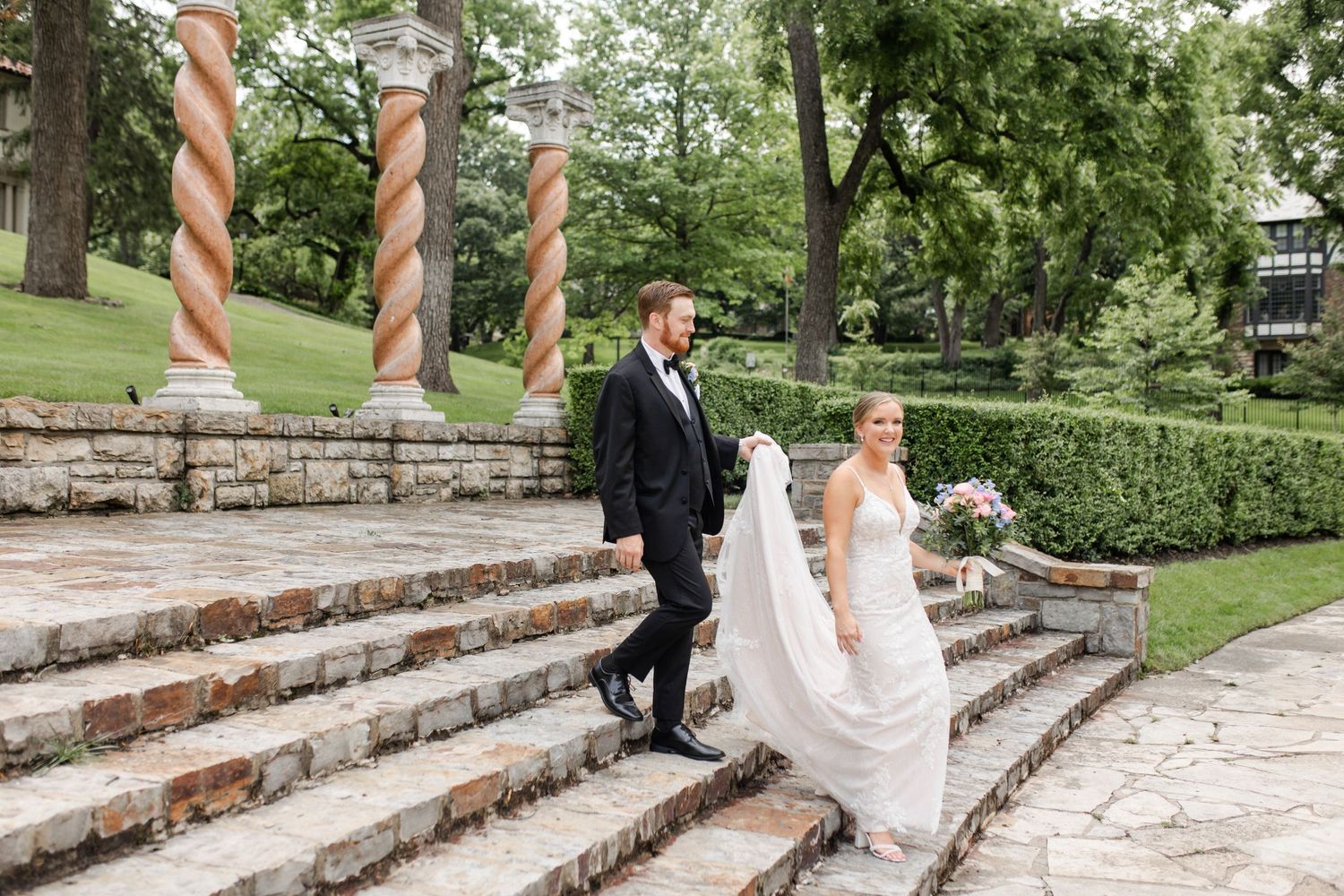A bride and groom descend stone steps in an elegant garden setting with decorative spiral columns in the background.