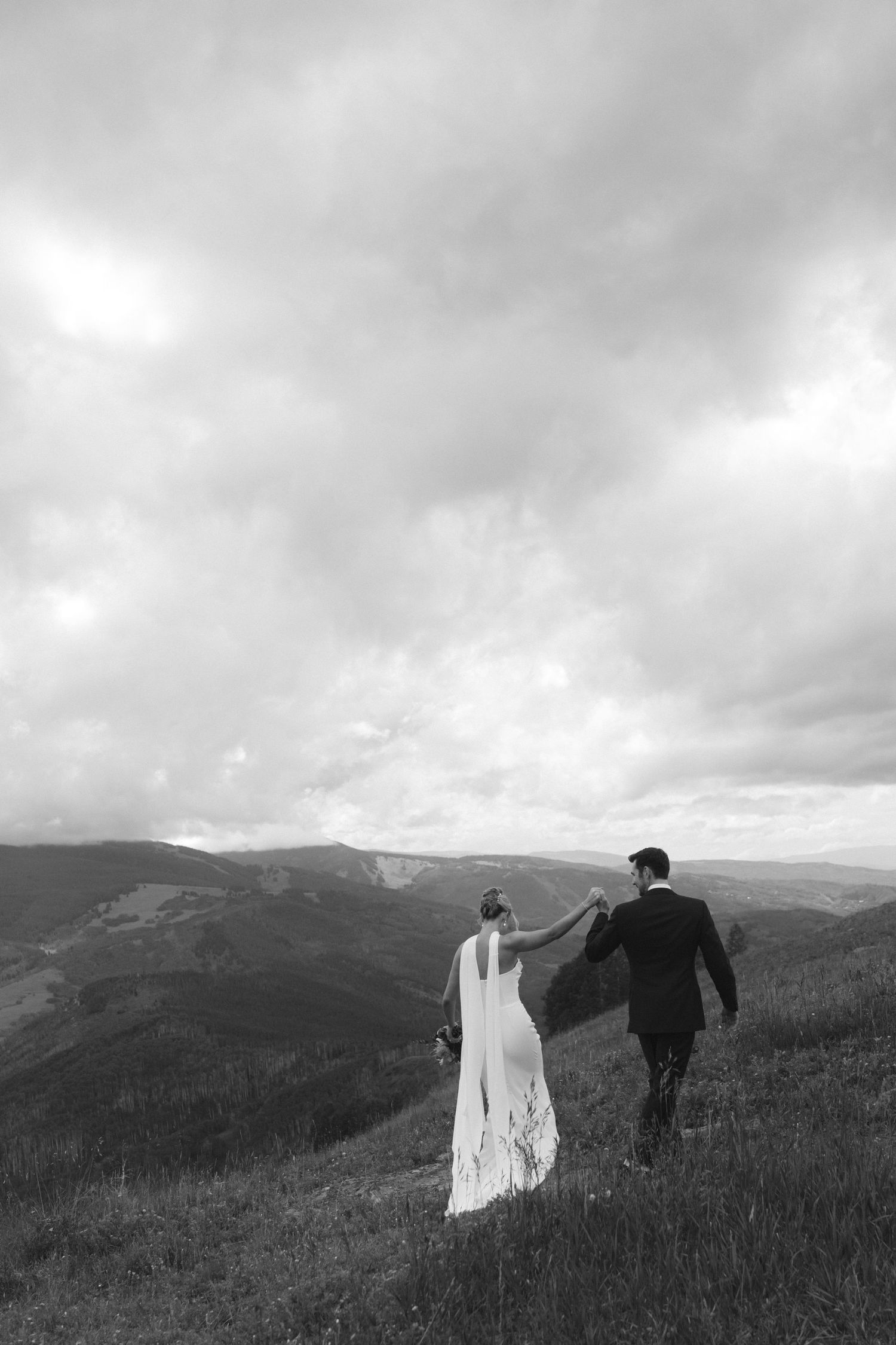 A black and white wedding photo capturing a couple walking hand in hand across a hilltop meadow with mountain views.