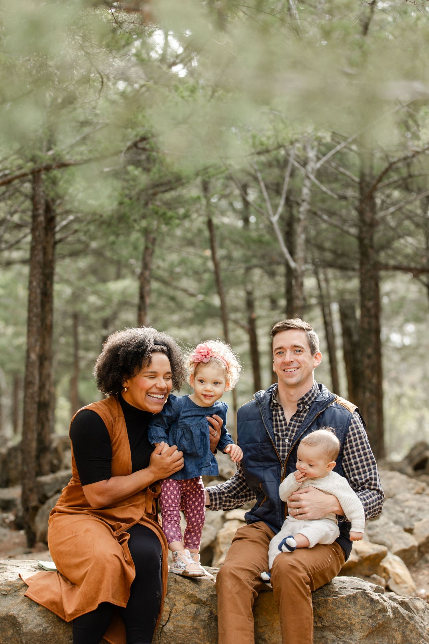 Family sitting together on rocks in pine forest during outdoor photo session at Shawnee Mission Park by Hannah Taylor.