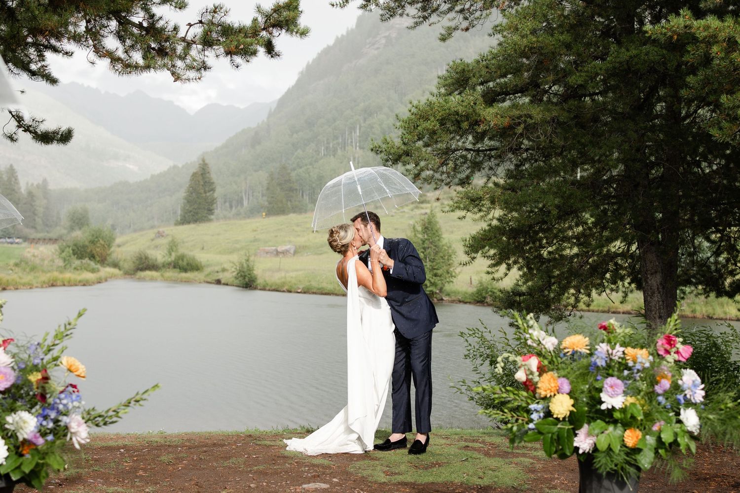 A couple shares an intimate moment under an umbrella by a misty lake surrounded by floral arrangements. Vail, Colorado Mountain Wedding.
