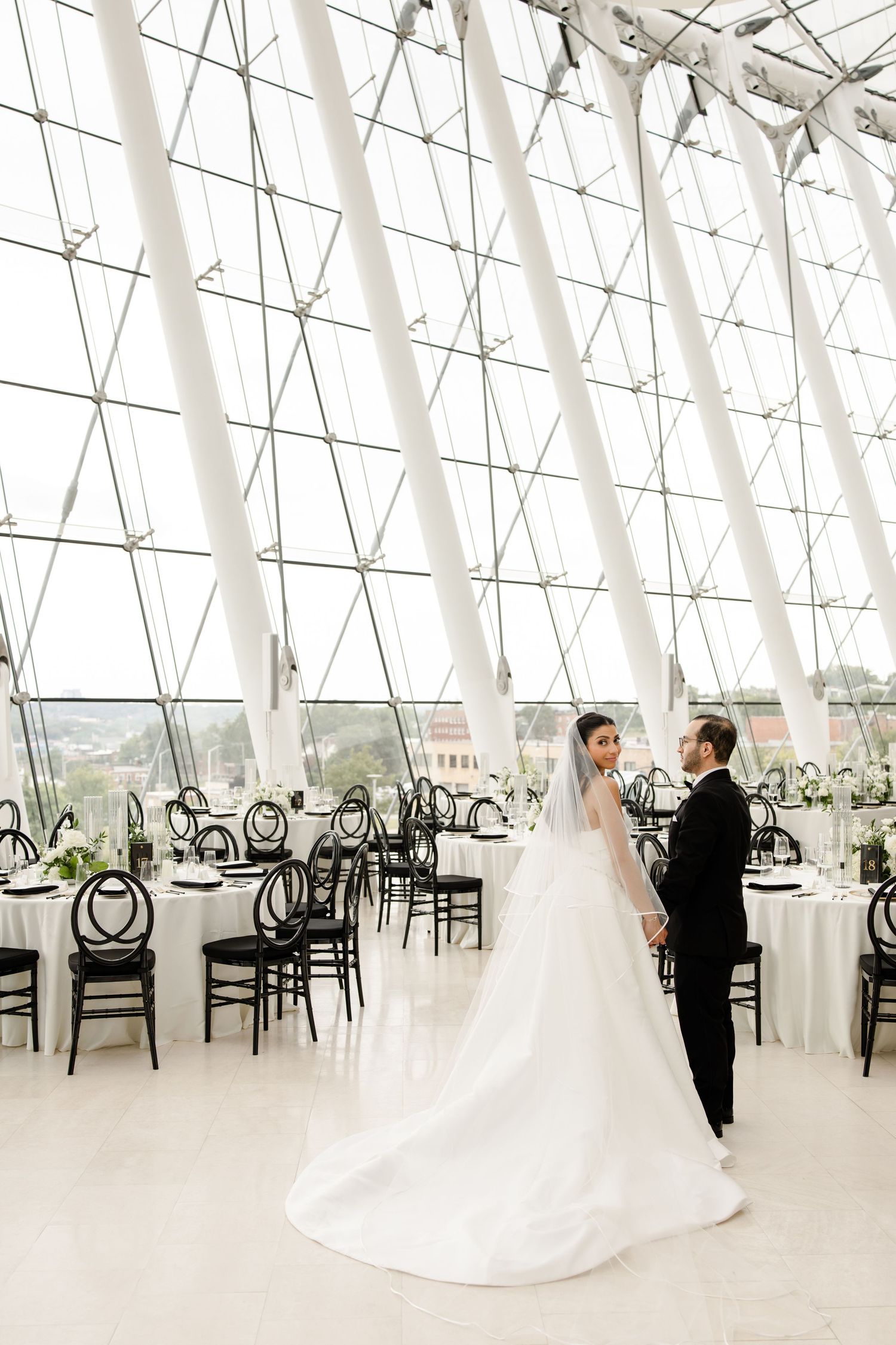 A wedding couple dances in a bright white reception venue with modern ghost chairs and elegant table settings.