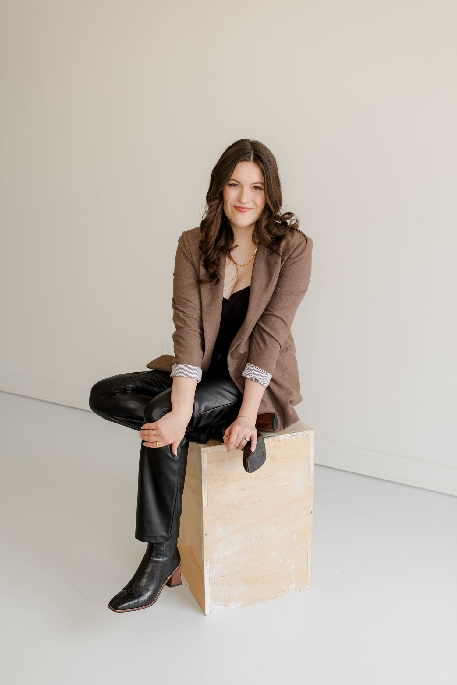 Modern fashion portrait showing brown blazer, leather pants and boots with minimalist wooden prop against white background.