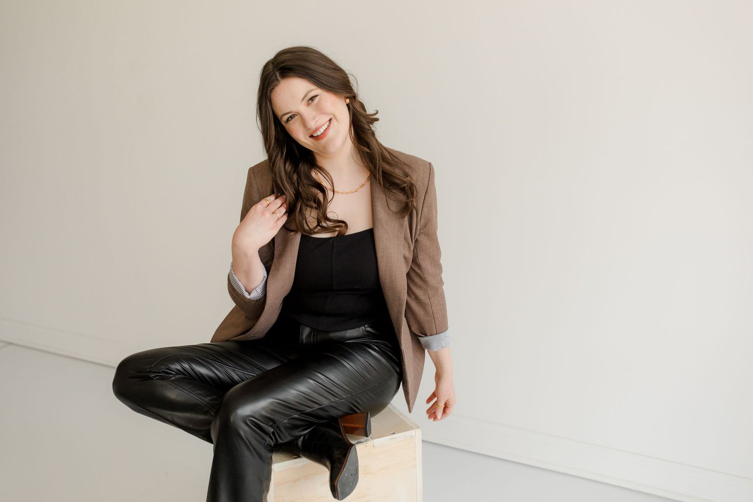 A woman wearing a brown blazer and black leather pants sits casually against a white wall, smiling and relaxed.