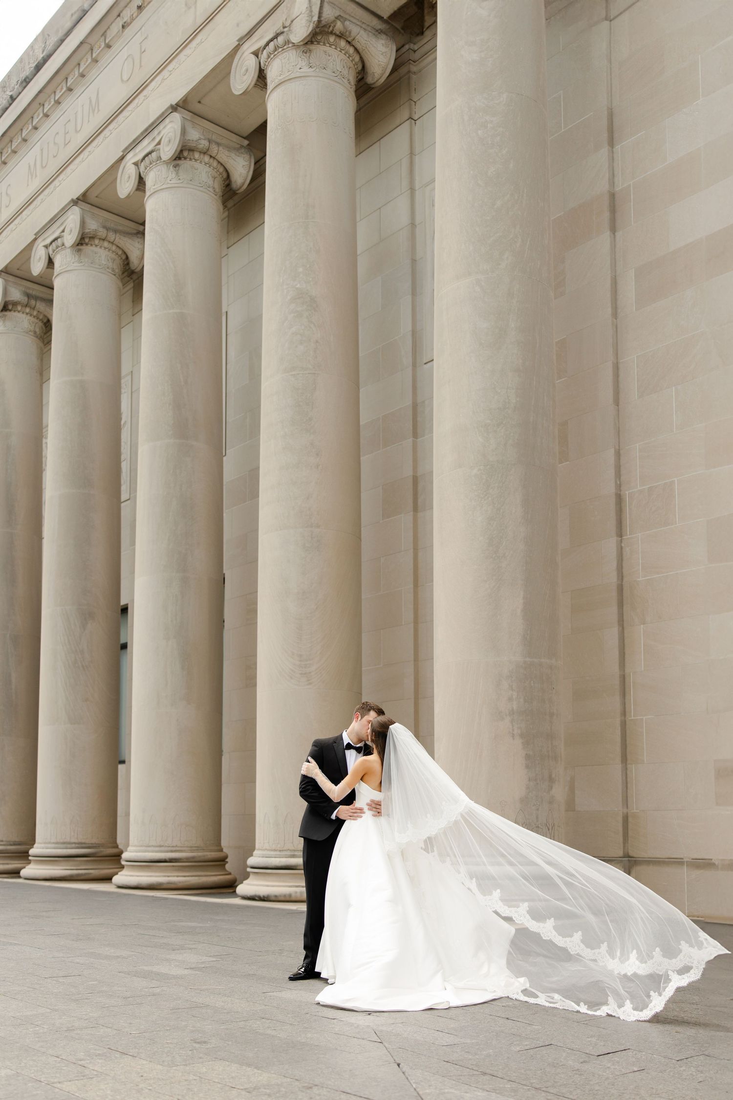 Elegant wedding couple embraces against a grand colonnade with flowing white veil creating romantic architectural backdrop.