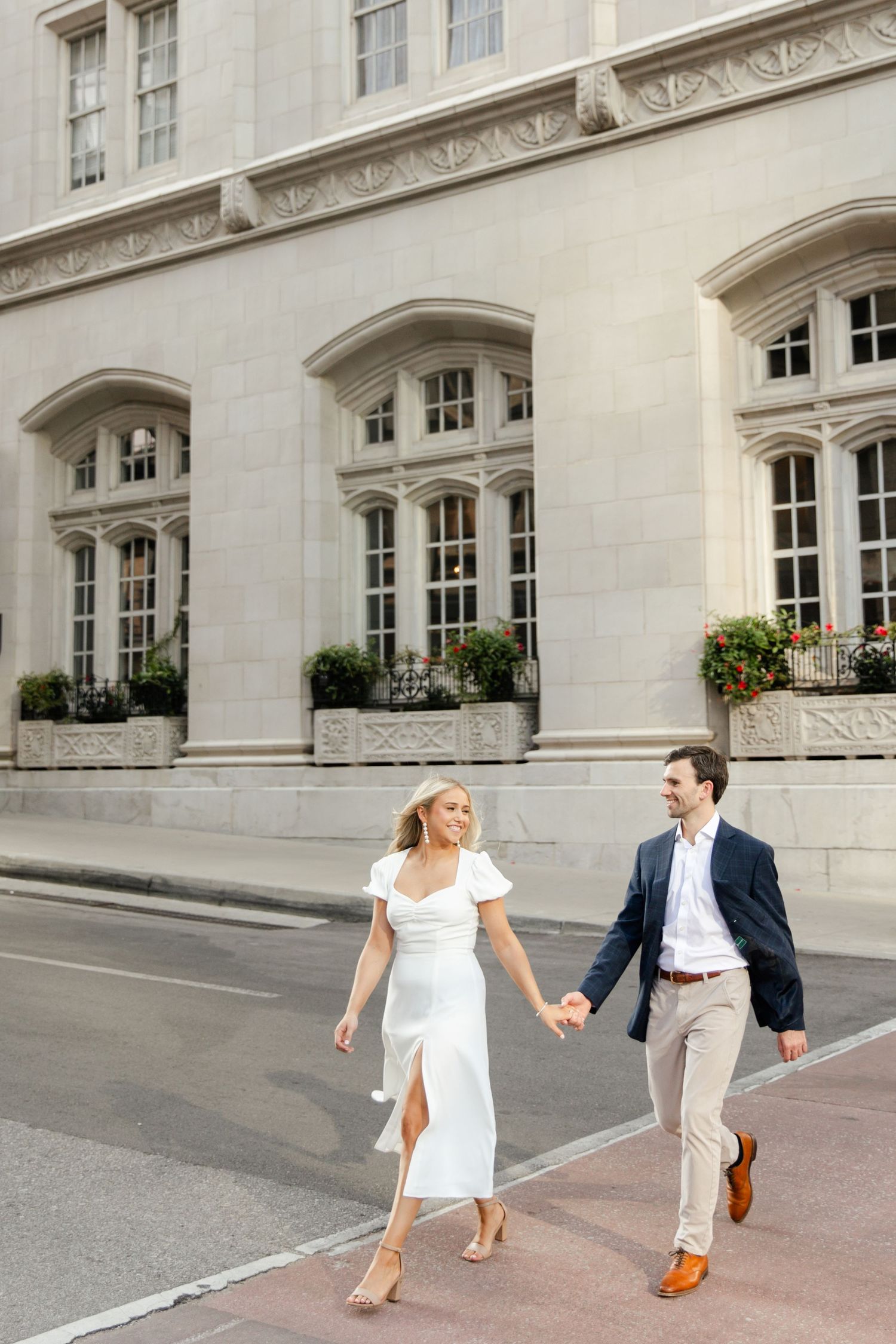 A couple strolls hand-in-hand past a grand building with ornate architecture and window boxes in front of Hotel Kansas City. City engagement