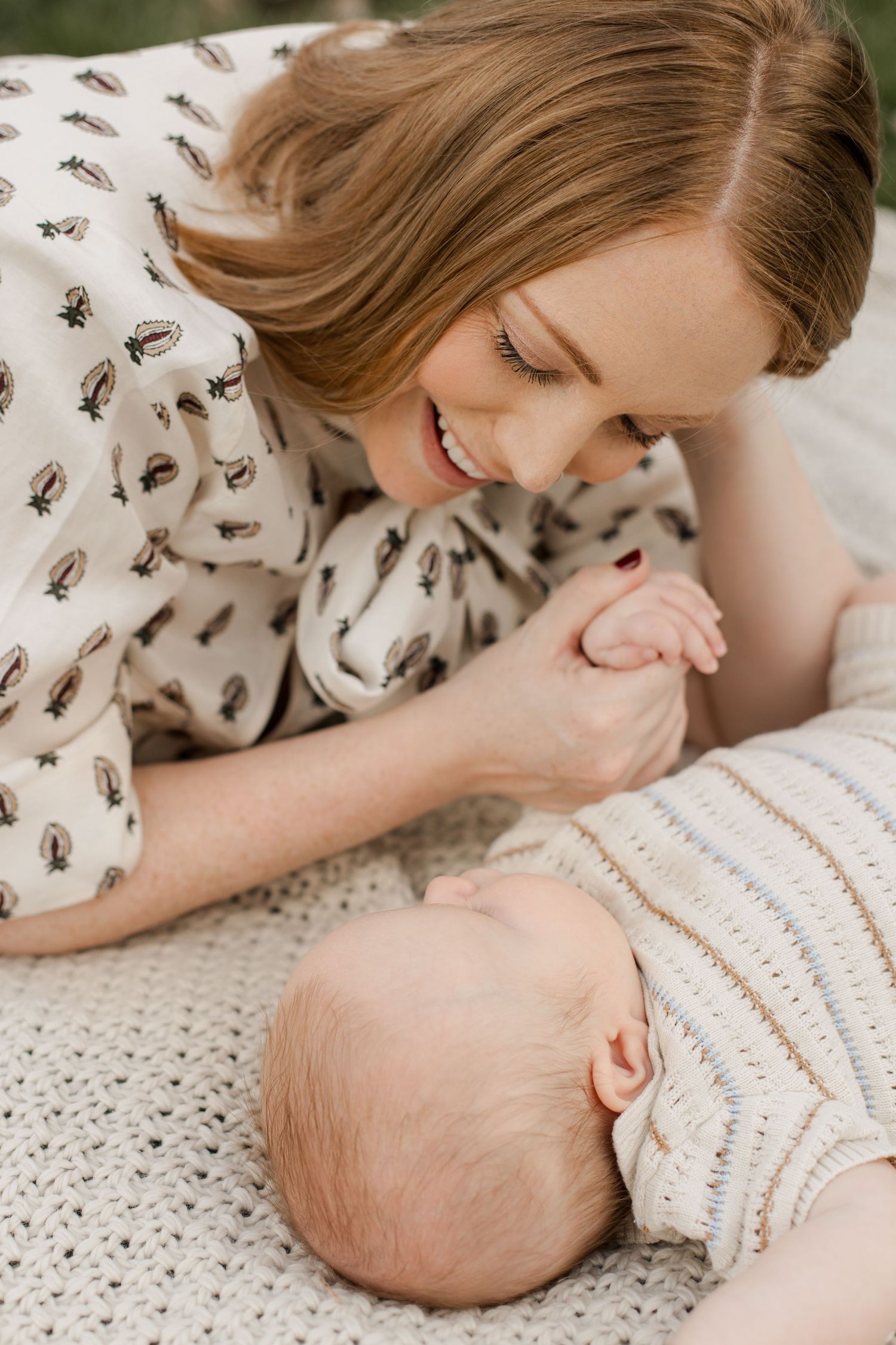 A tender moment as a mother in a patterned dress leans over to connect with her sleeping baby on a knit blanket.