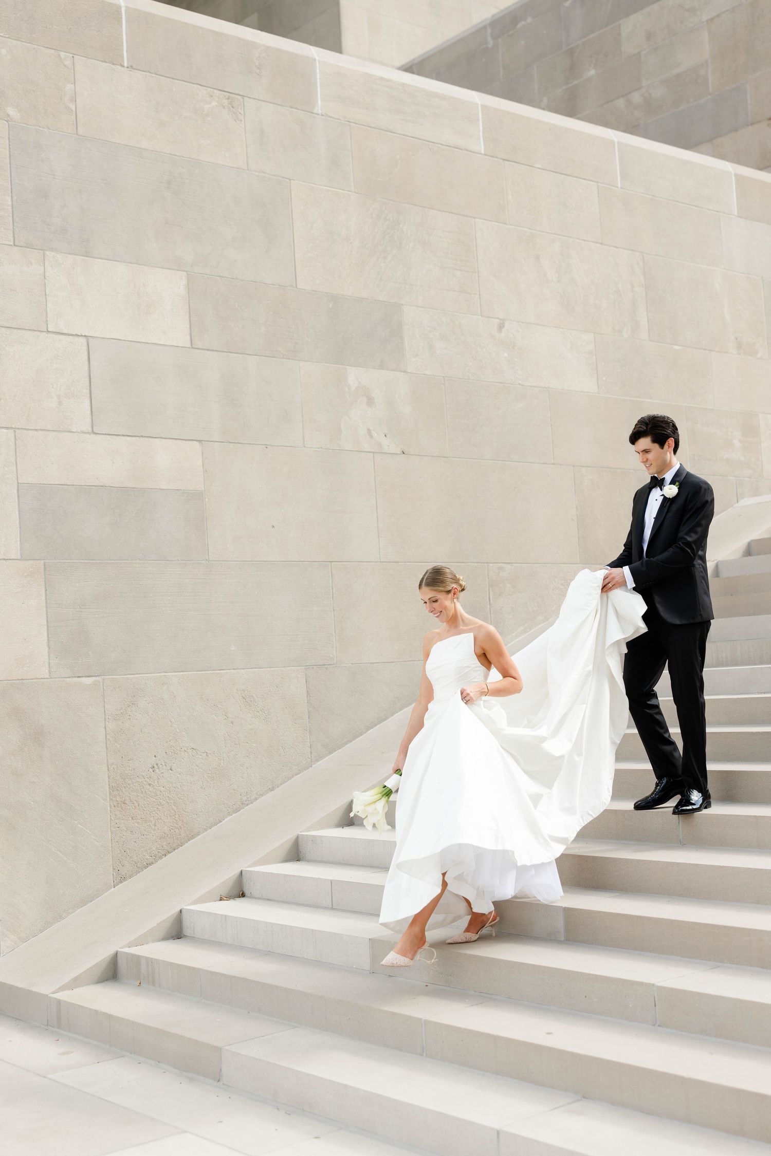 A bride in a flowing white dress gracefully descends stone steps while her groom assists with her train.
