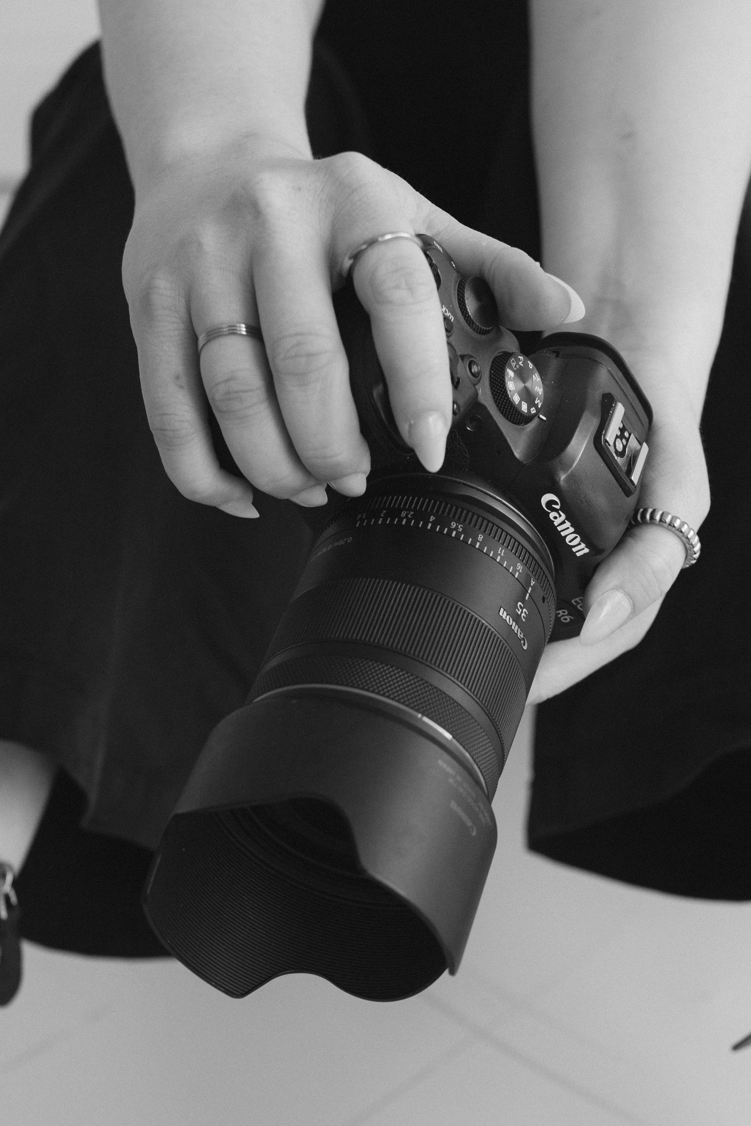 Close-up black and white photo of hands holding a Canon camera with telephoto lens against light background.