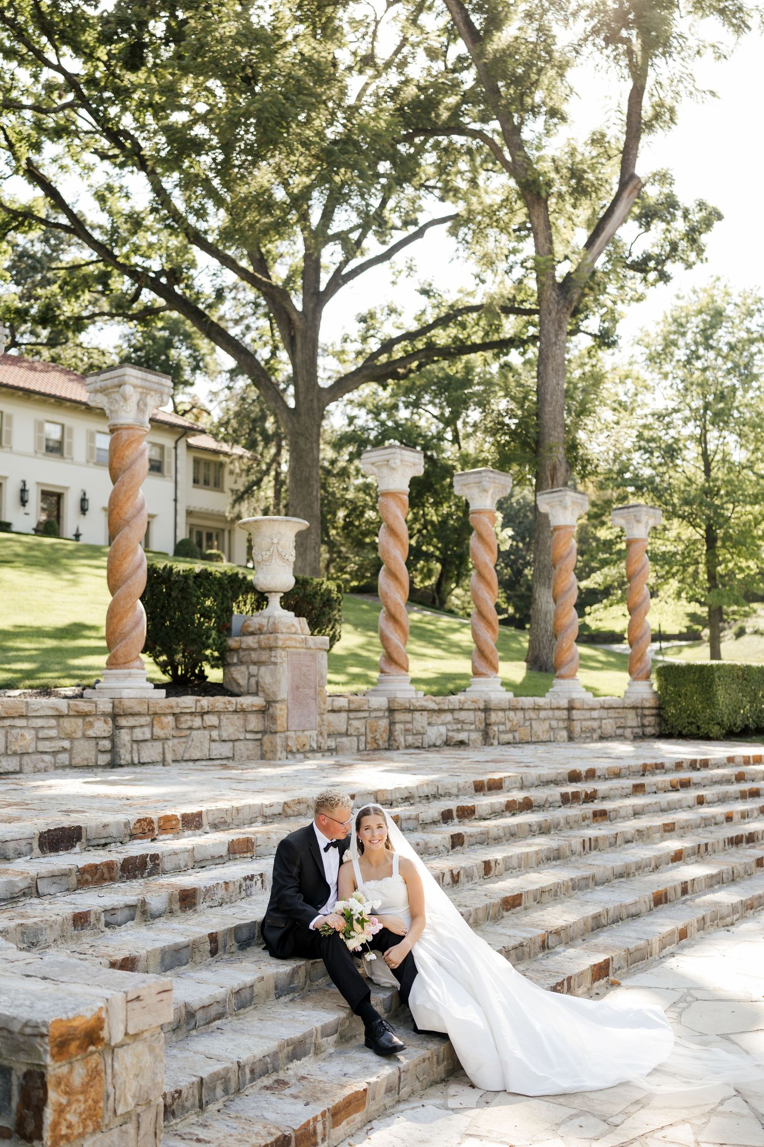 A couple in wedding attire poses romantically on elegant stone steps with decorative pillars and trees in the background.