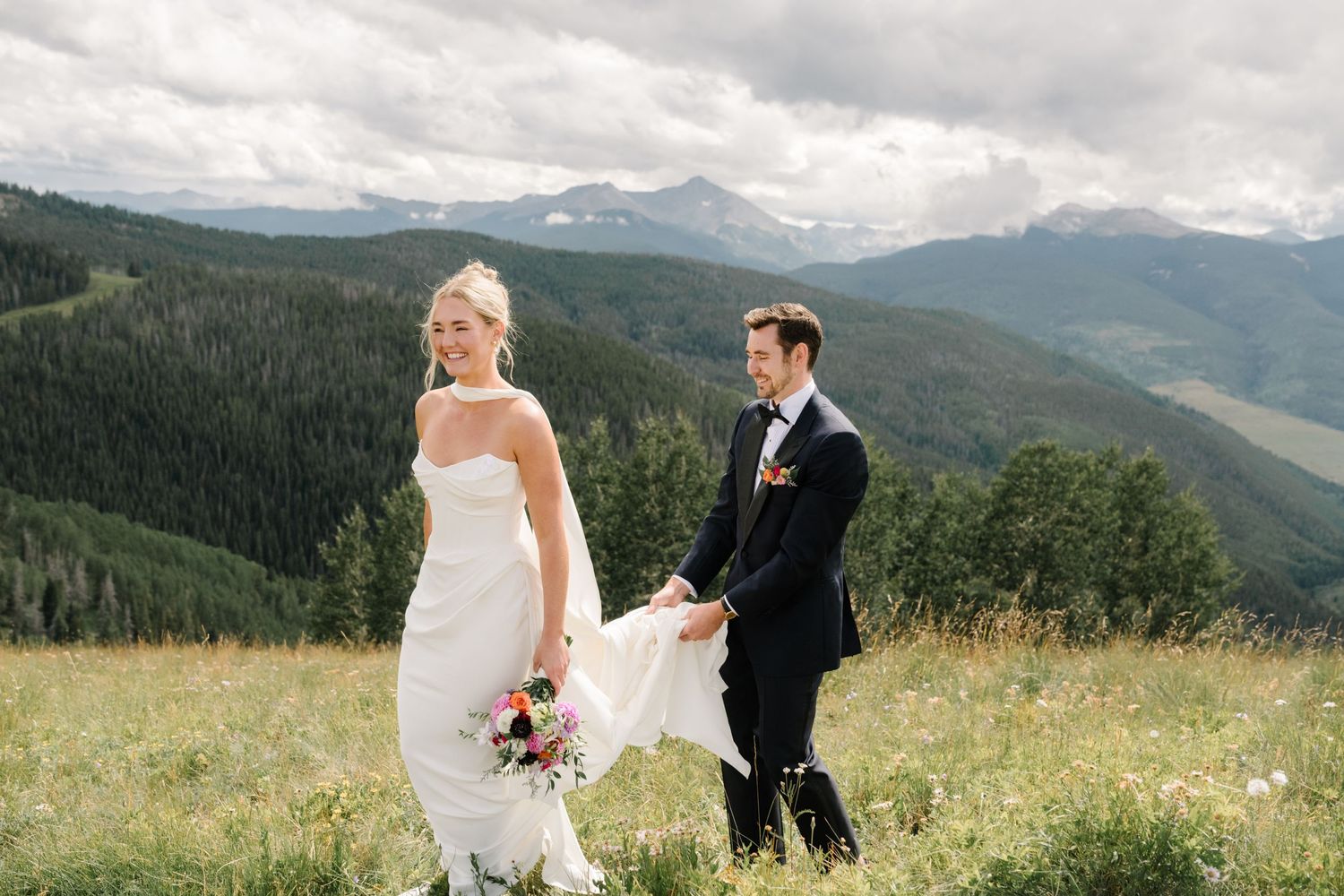 A couple in wedding attire walks across a mountain meadow with dramatic mountain peaks and forest views in Colorado.
