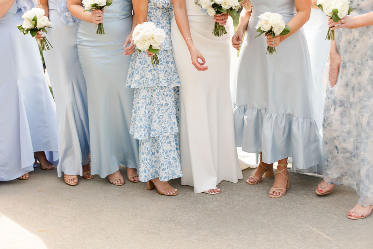 Wedding party members in flowing light blue and floral dresses hold white bouquets while standing together.