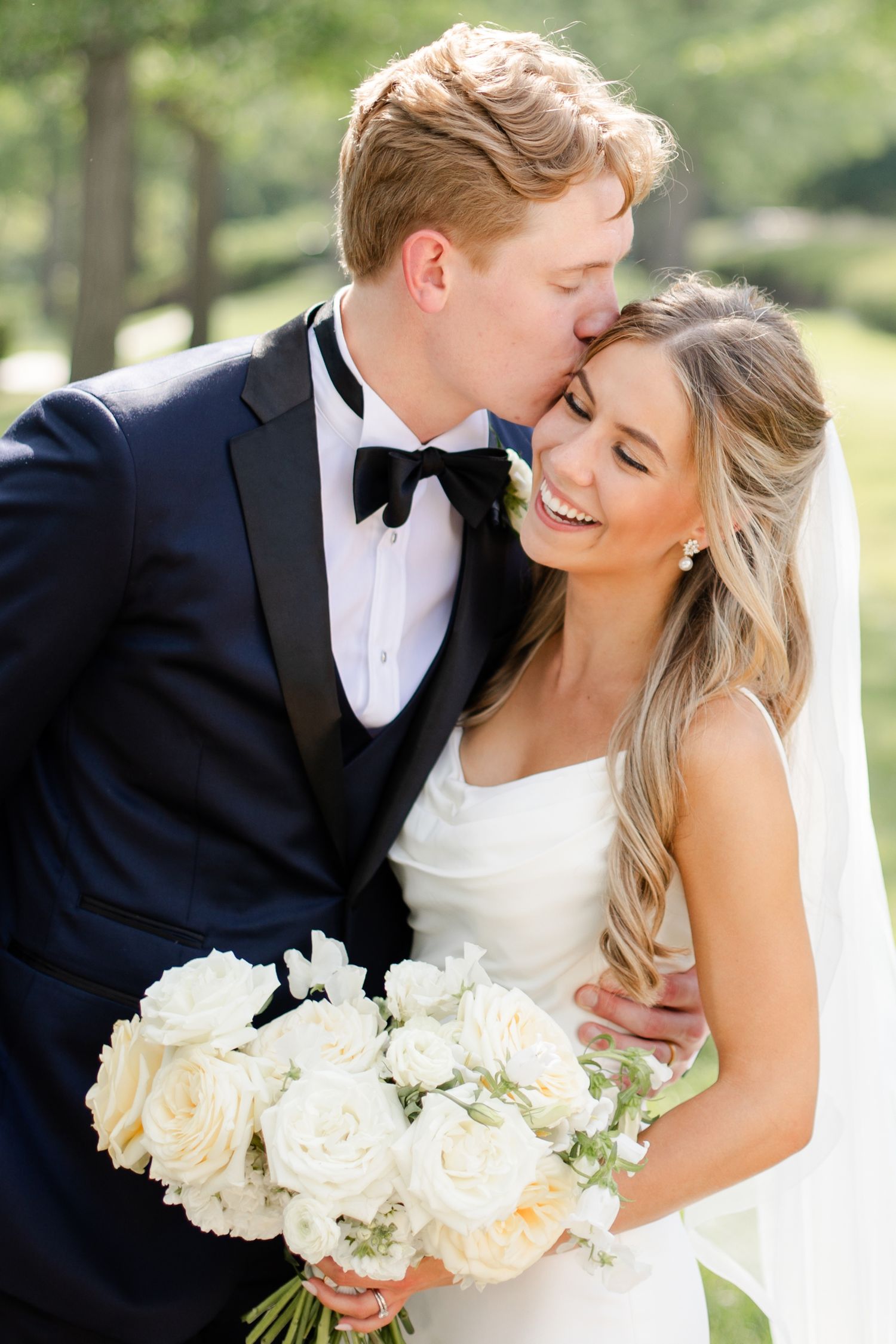 Romantic wedding portrait with couple sharing intimate moment among trees holding white bouquet.