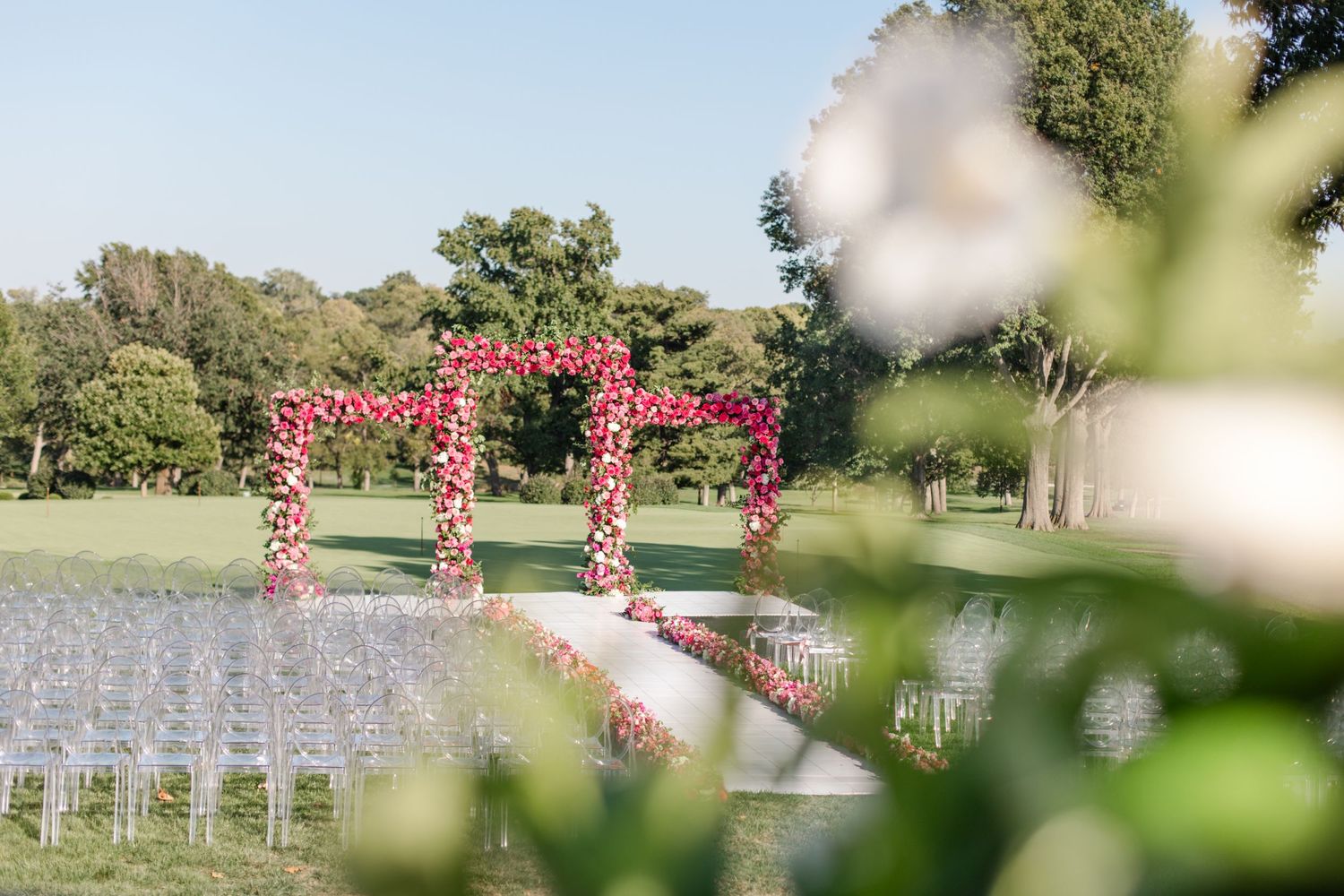 Outdoor wedding ceremony setup with pink floral arch and white chairs on a sunny day at Kansas City Country Club.