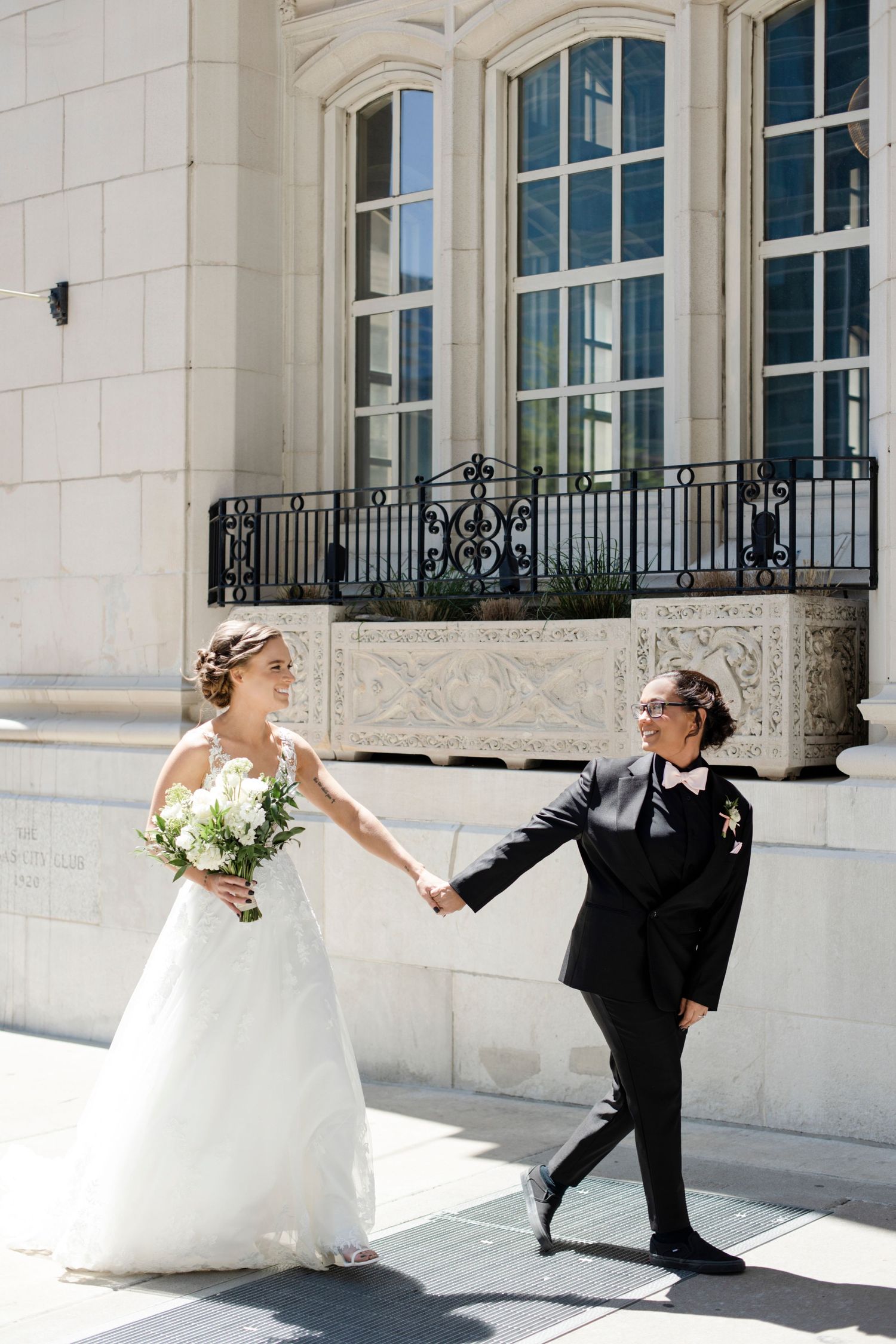 Wedding couple playfully poses in front of an elegant white building with ornate windows. Hotel Kansas City. LGBTQ Friendly wedding photos.