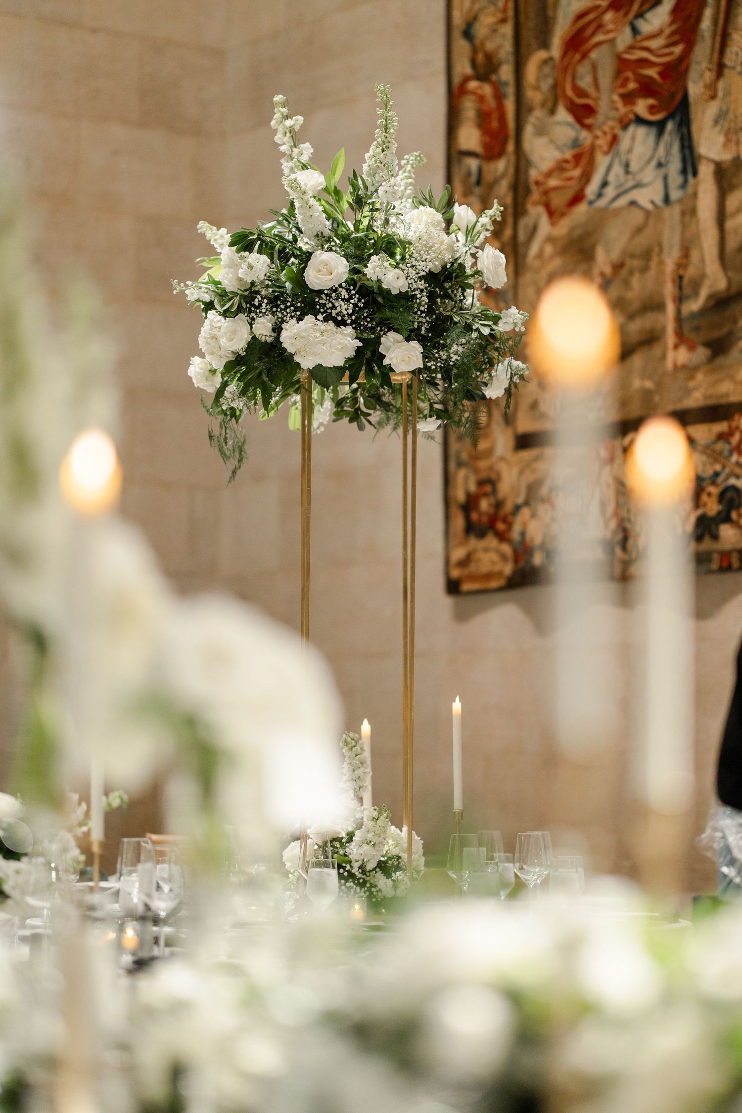 A tall white floral centerpiece on a gold stand with glowing candles and tapestries in the background at their Art Museum Wedding in Kansas.