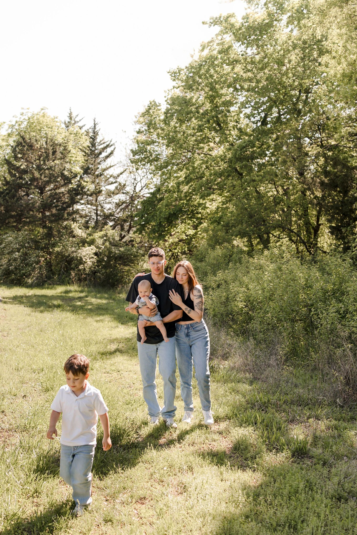 A family walks together on a grassy trail surrounded by lush green trees on a sunny summer day.
