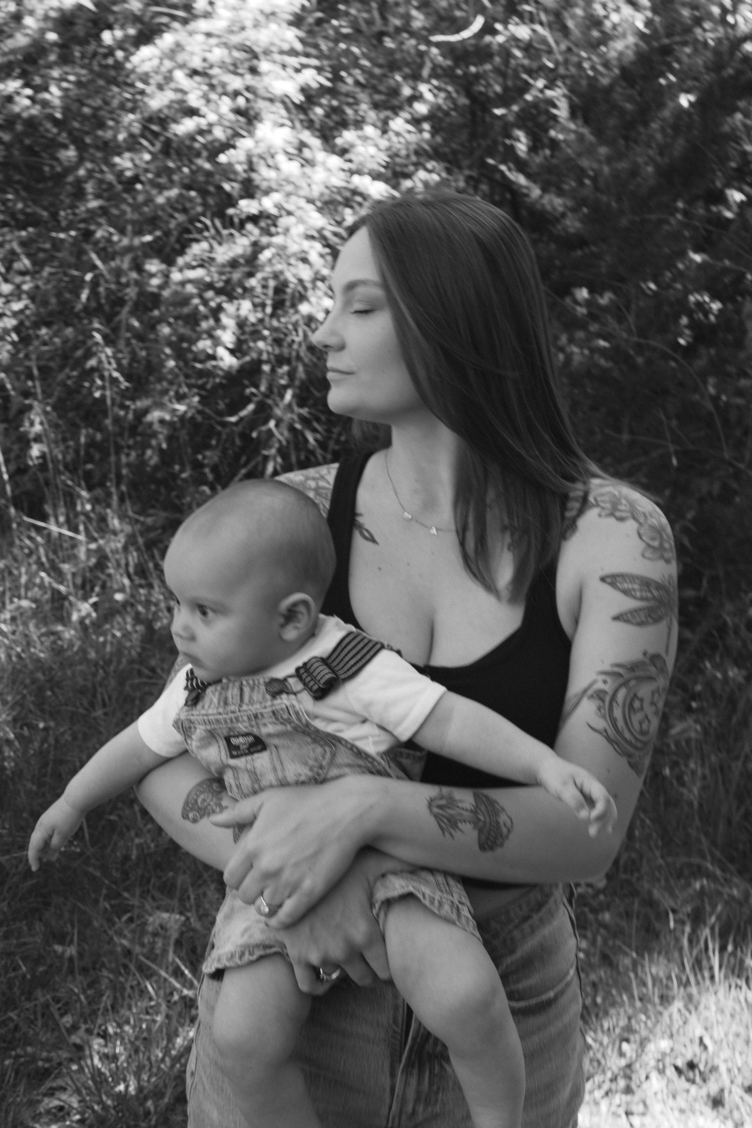 A black and white portrait of a mother embracing her baby outdoors in a natural setting.