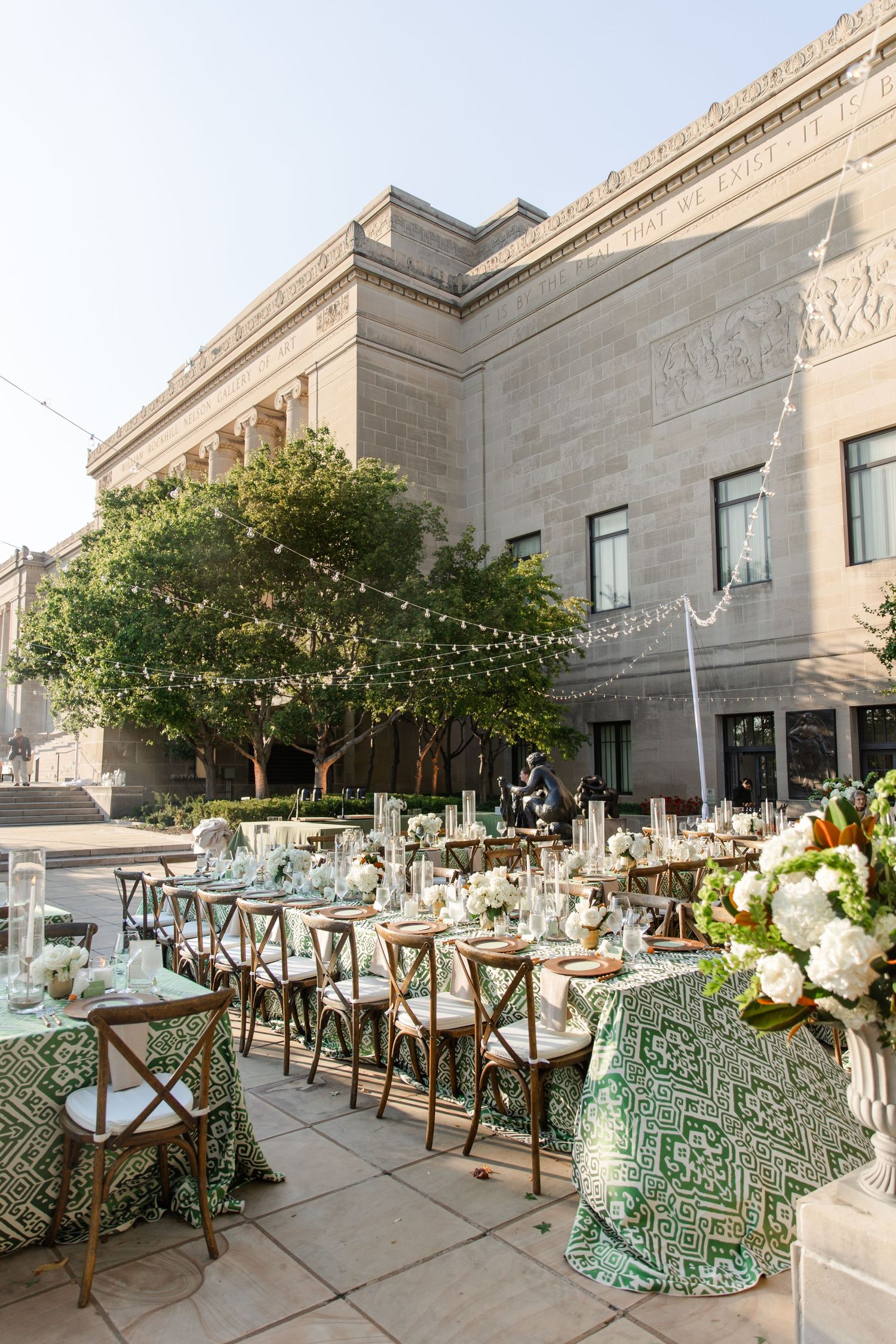An elegant outdoor wedding reception setup with green tablecloths and cross-back chairs against a stone building. Nelson Atkins Art Museum.