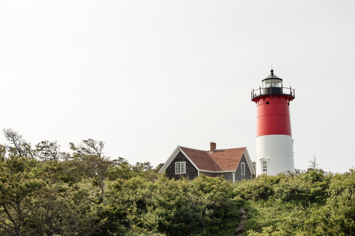 Red and white lighthouse rising above lush green trees beside a charming cottage on Cape Cod, Massachusetts.