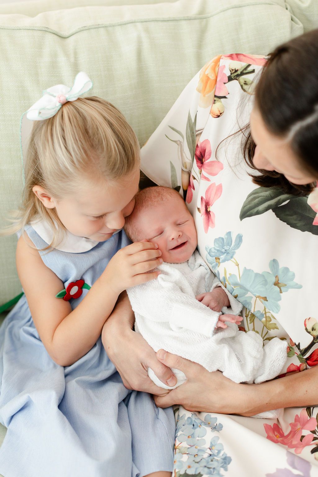 A young girl in a blue dress gives a tender kiss to a newborn baby being held in floral patterned fabric.