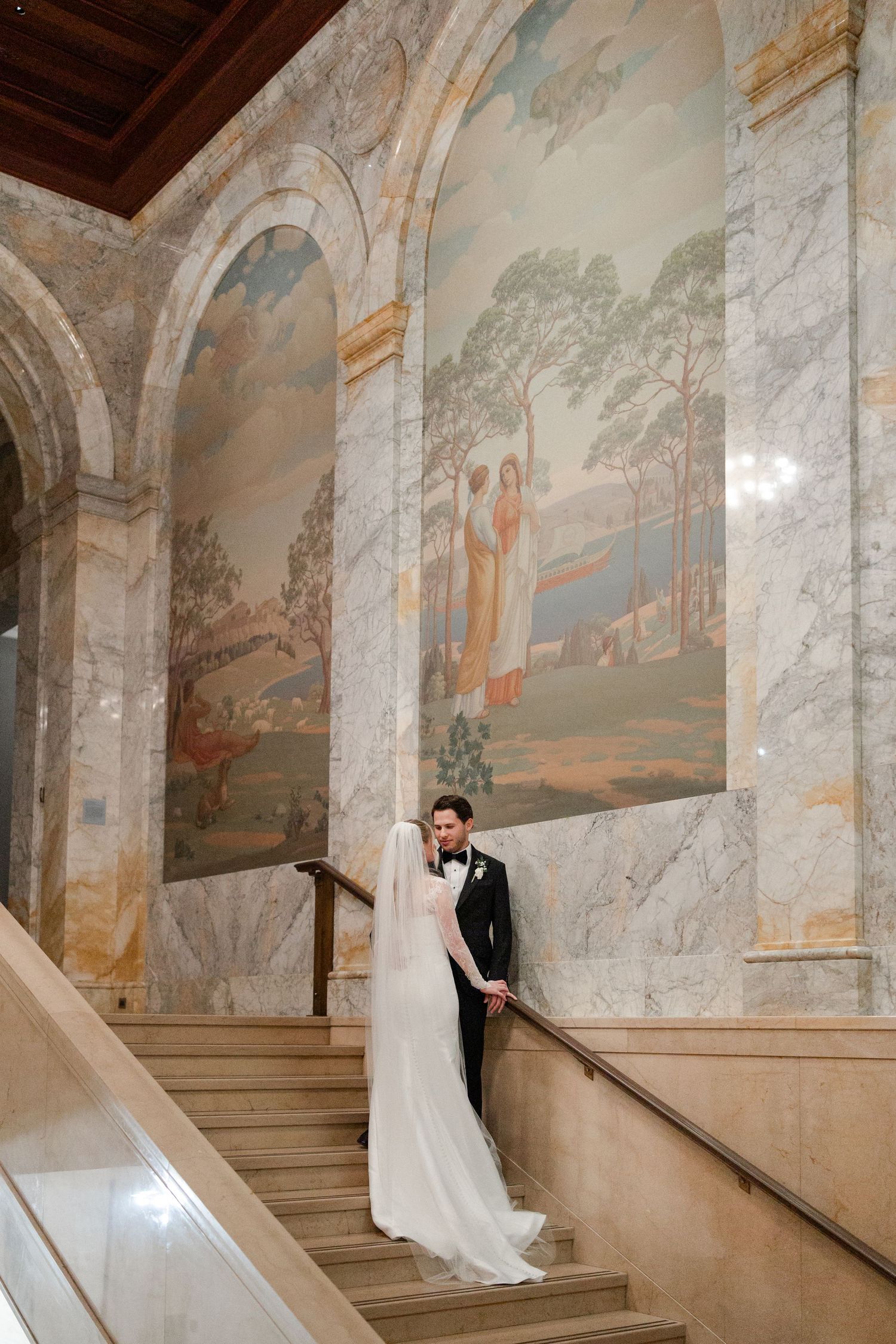 Wedding couple poses on grand marble staircase with ornate wall murals at Nelson Atkins Art Museum in Kansas City.