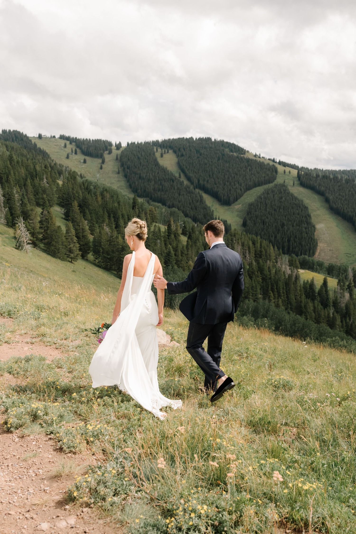 A couple in wedding attire walks along a mountain trail with ski slopes and evergreen forests in the background.