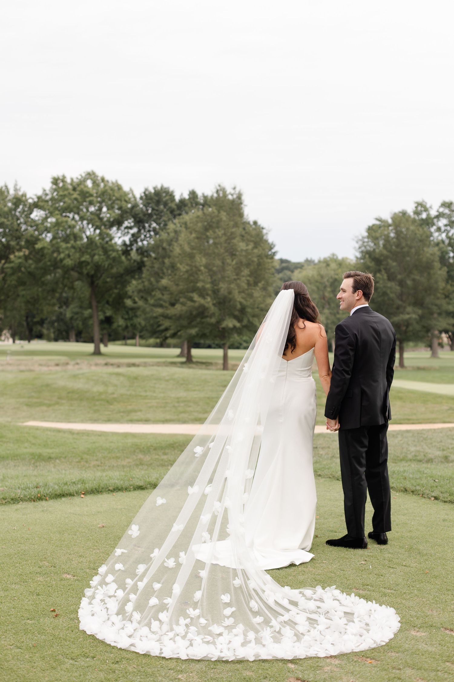 A couple in wedding attire stands on a lush golf course with a long floral-adorned veil trailing behind on the grass.