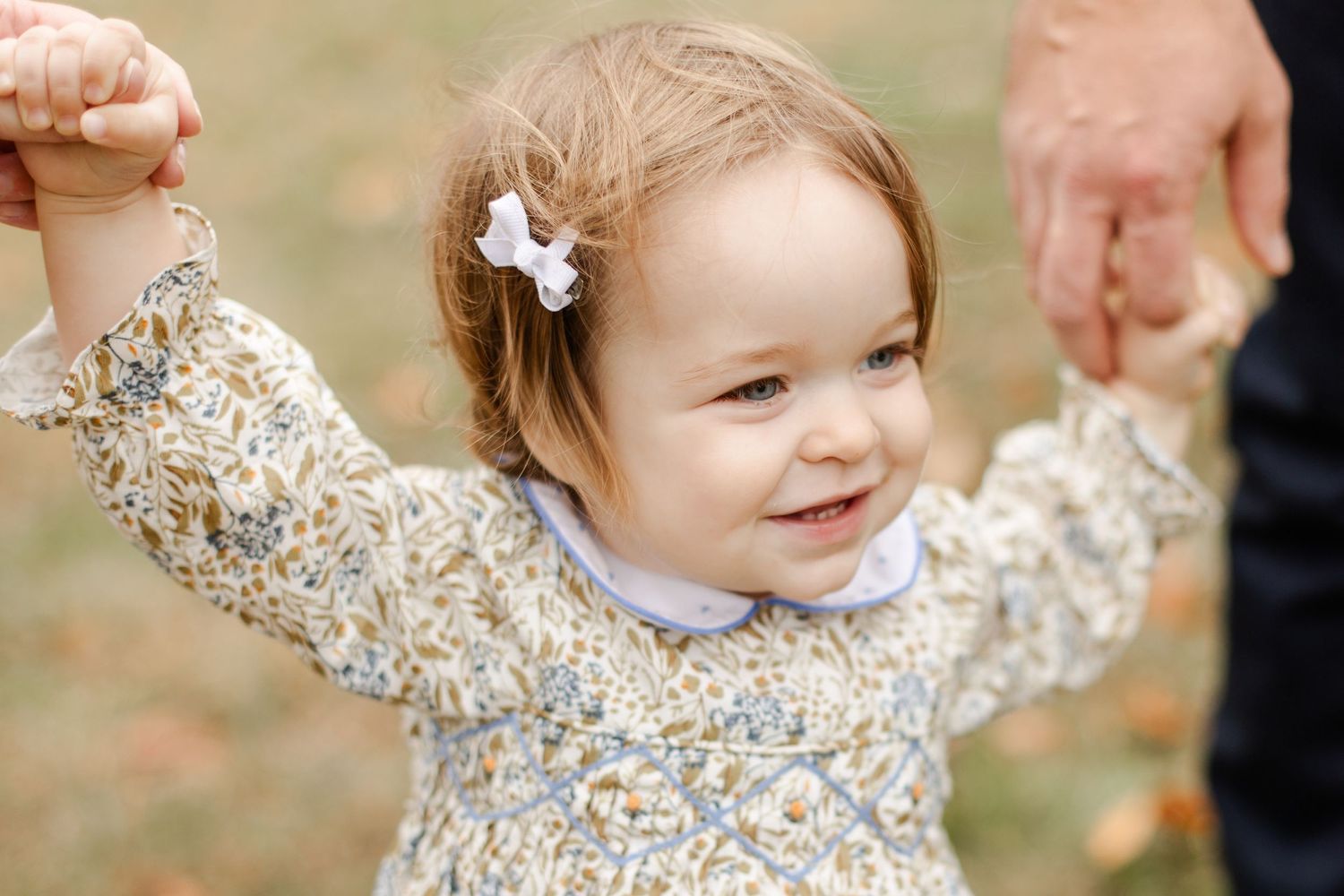 Young child wearing floral dress smiles while holding hands and walking outdoors.