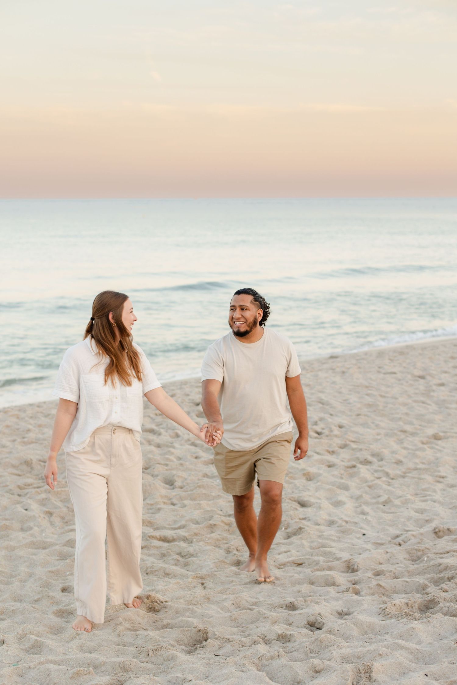 Two people stroll together on a sandy beach at dusk wearing light colored clothing. Florida wedding photographer Hannah Rogg Taylor.