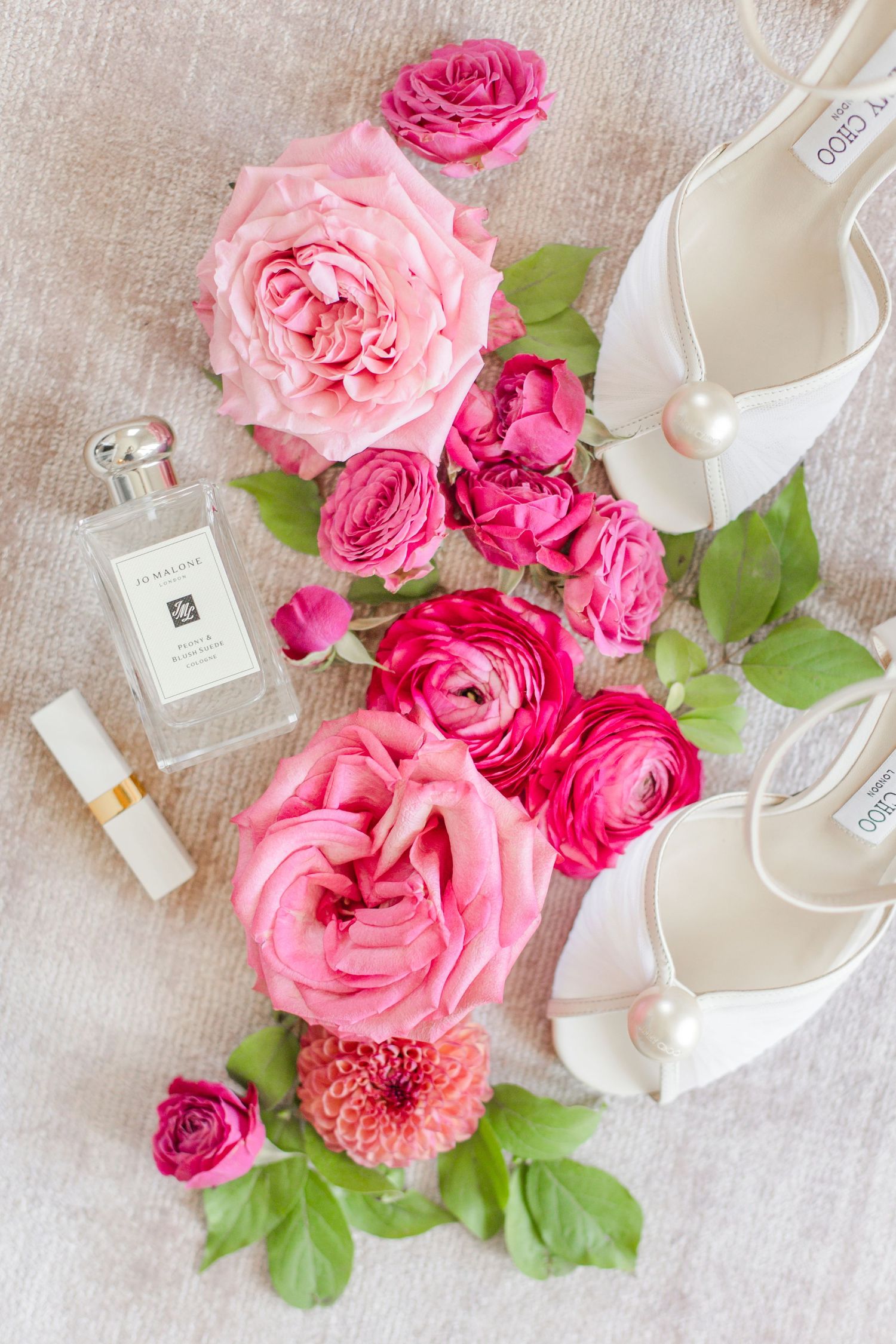 Elegant white bridal shoes with pink roses, perfume bottle and green leaves arranged on light fabric background.