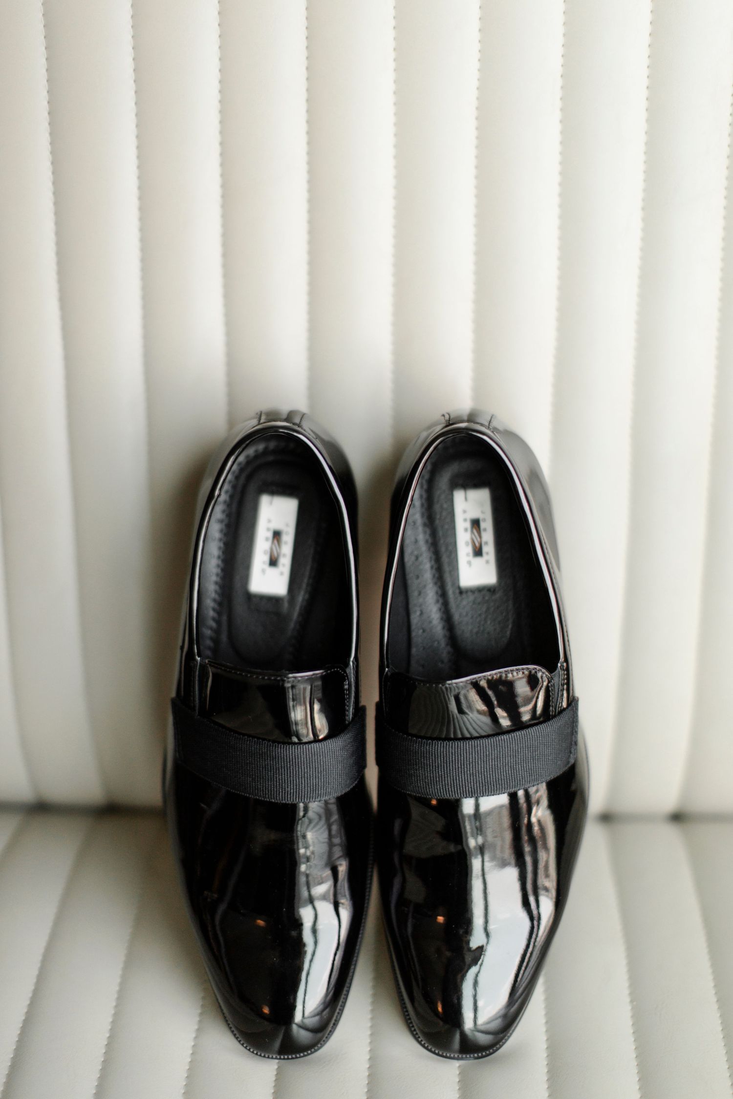 A pair of shiny black patent leather formal dress shoes with elastic straps sits against a white striped background.